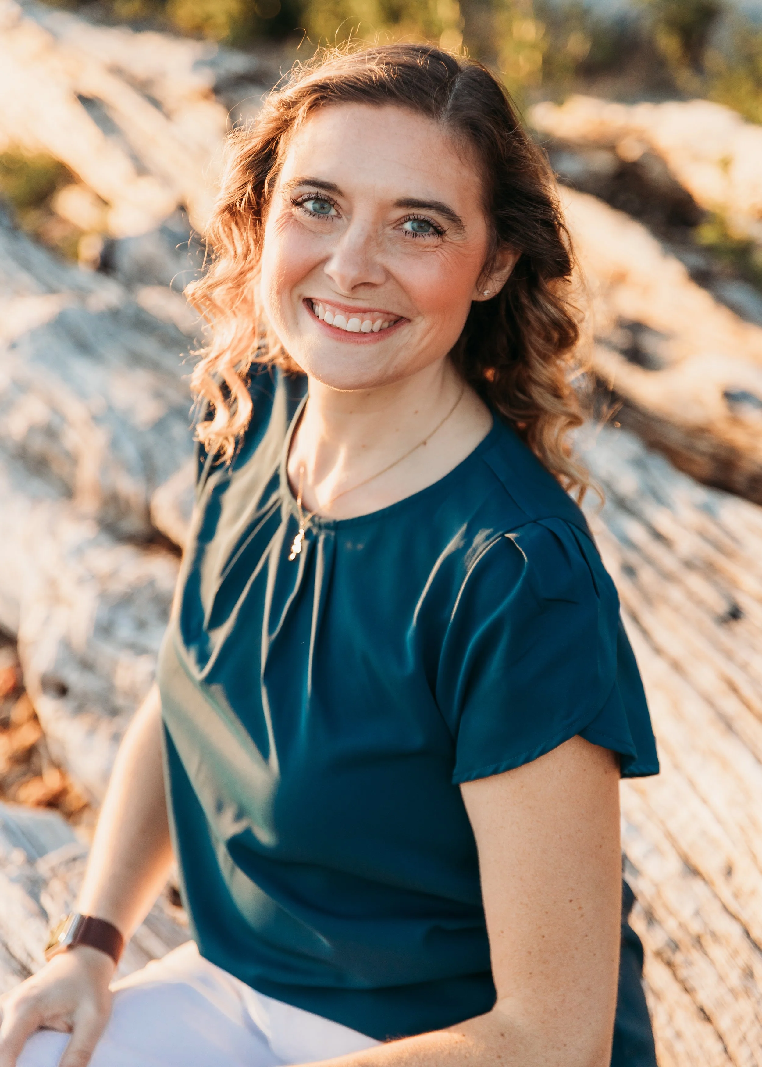 Close-up image of a smiling woman with blue eyes and curly hair, wearing a dark blue blouse, outdoors with trees and a rocky background, captured during golden hour lighting.