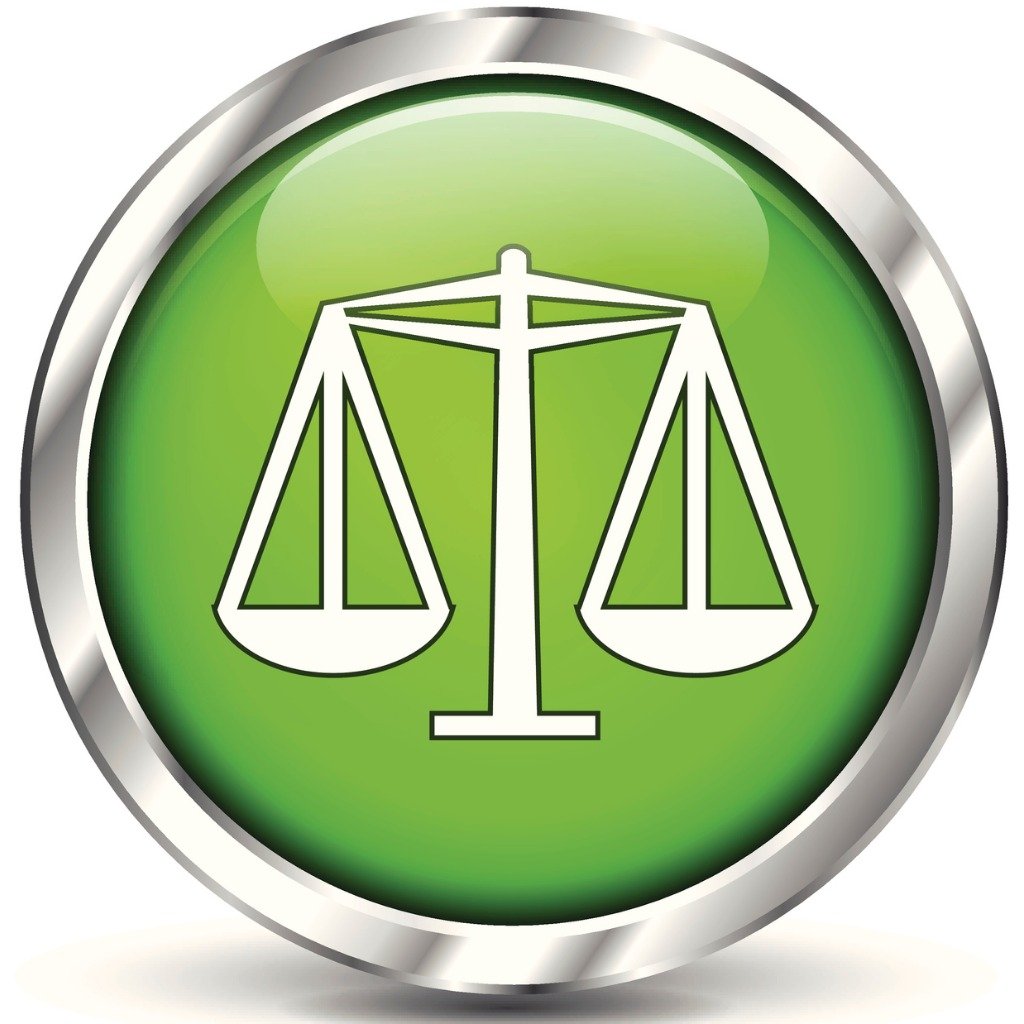 A circular button with a silver rim and a green background, featuring a white outline of a balanced scale symbol in the center.