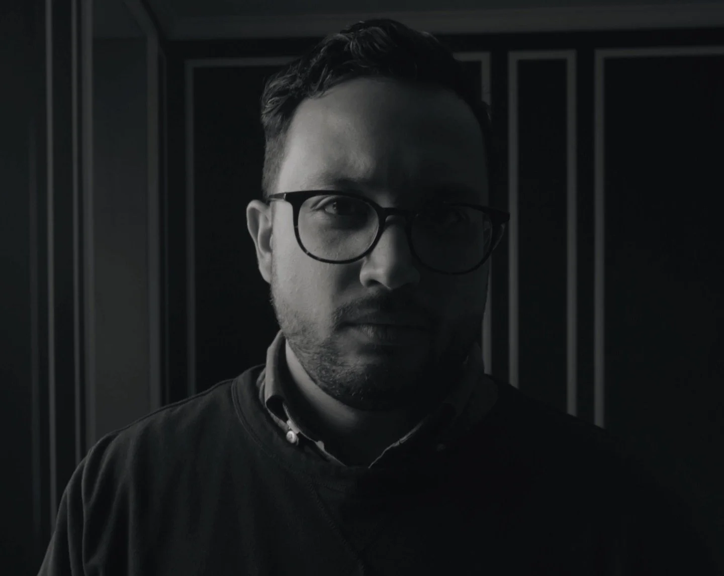 Black and white portrait of a man with glasses, dark background, serious expression, wearing a jacket.