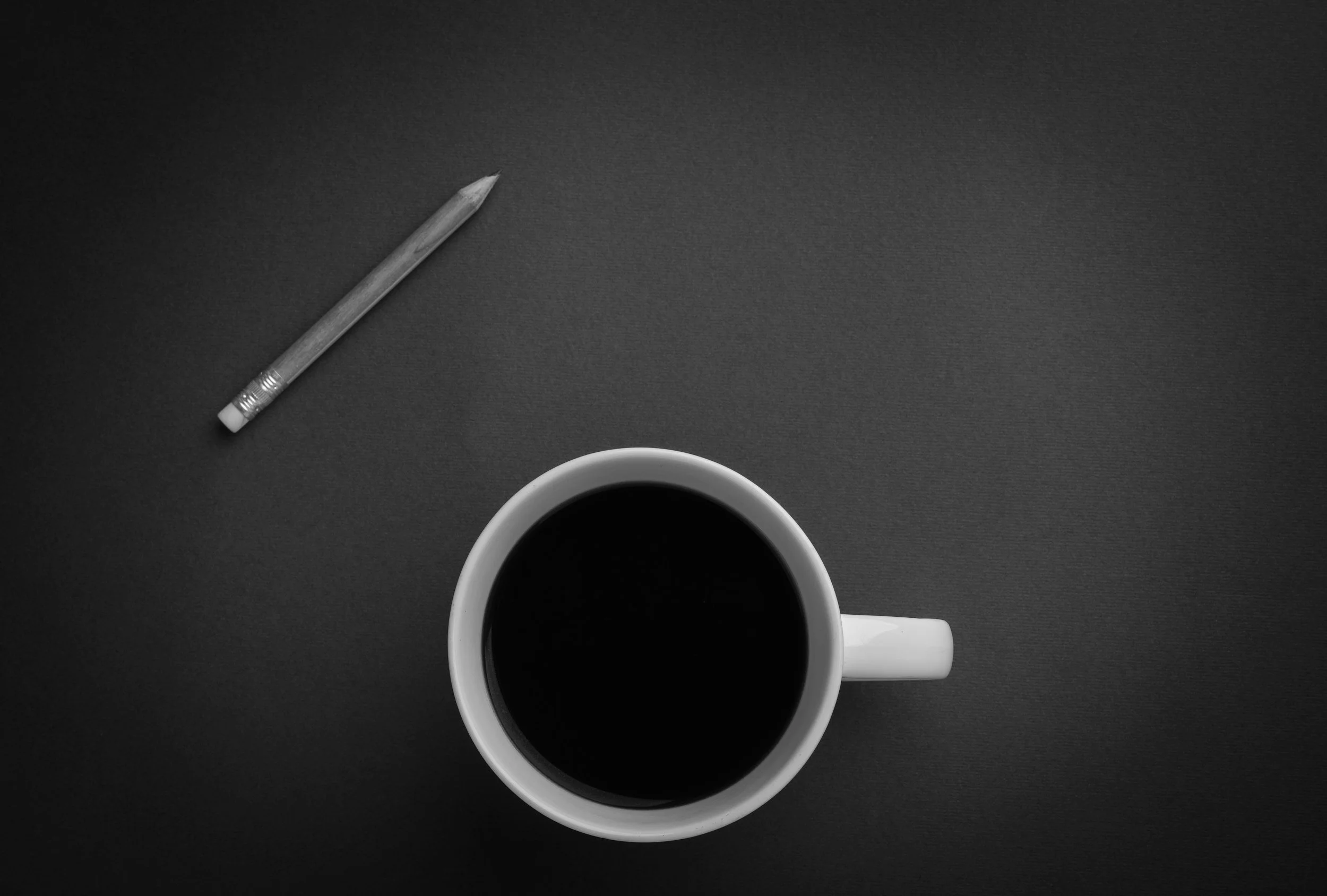 A top-down view of a white coffee mug filled with black coffee, placed on a dark gray surface, with a pencil to the left.