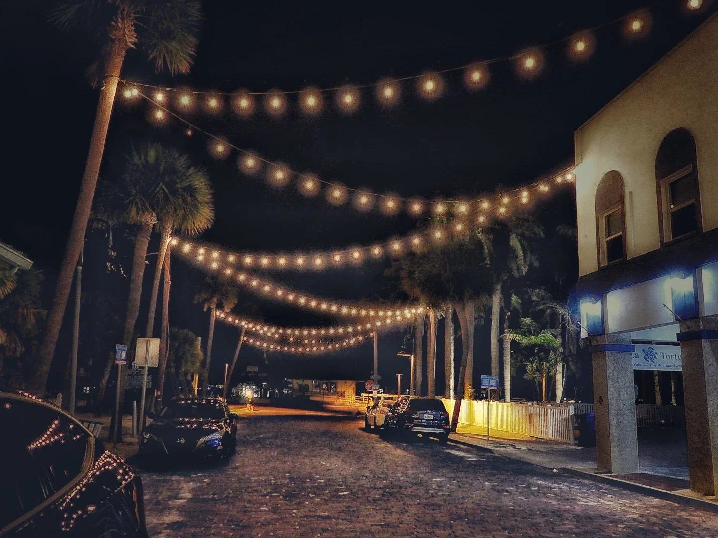 Night scene of a cobblestone street with parked cars, illuminated by string lights overhead. Palm trees line the street and buildings are visible on the right, with some lit windows and signs.