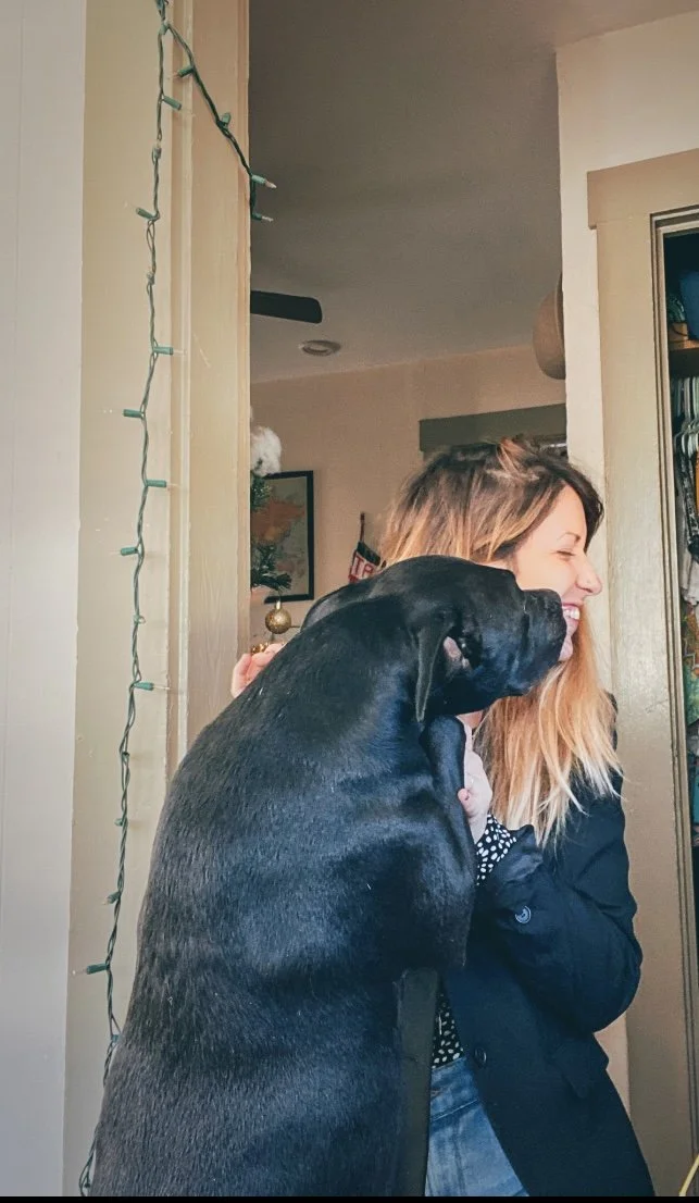 A woman with long blonde hair smiling while hugging a large black and gray dog inside a home, with Christmas decorations in the background.