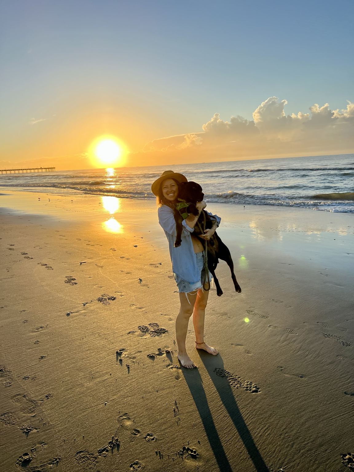 A woman holding a black dog on a beach during sunset, with footprints in the sand and clouds in the sky.