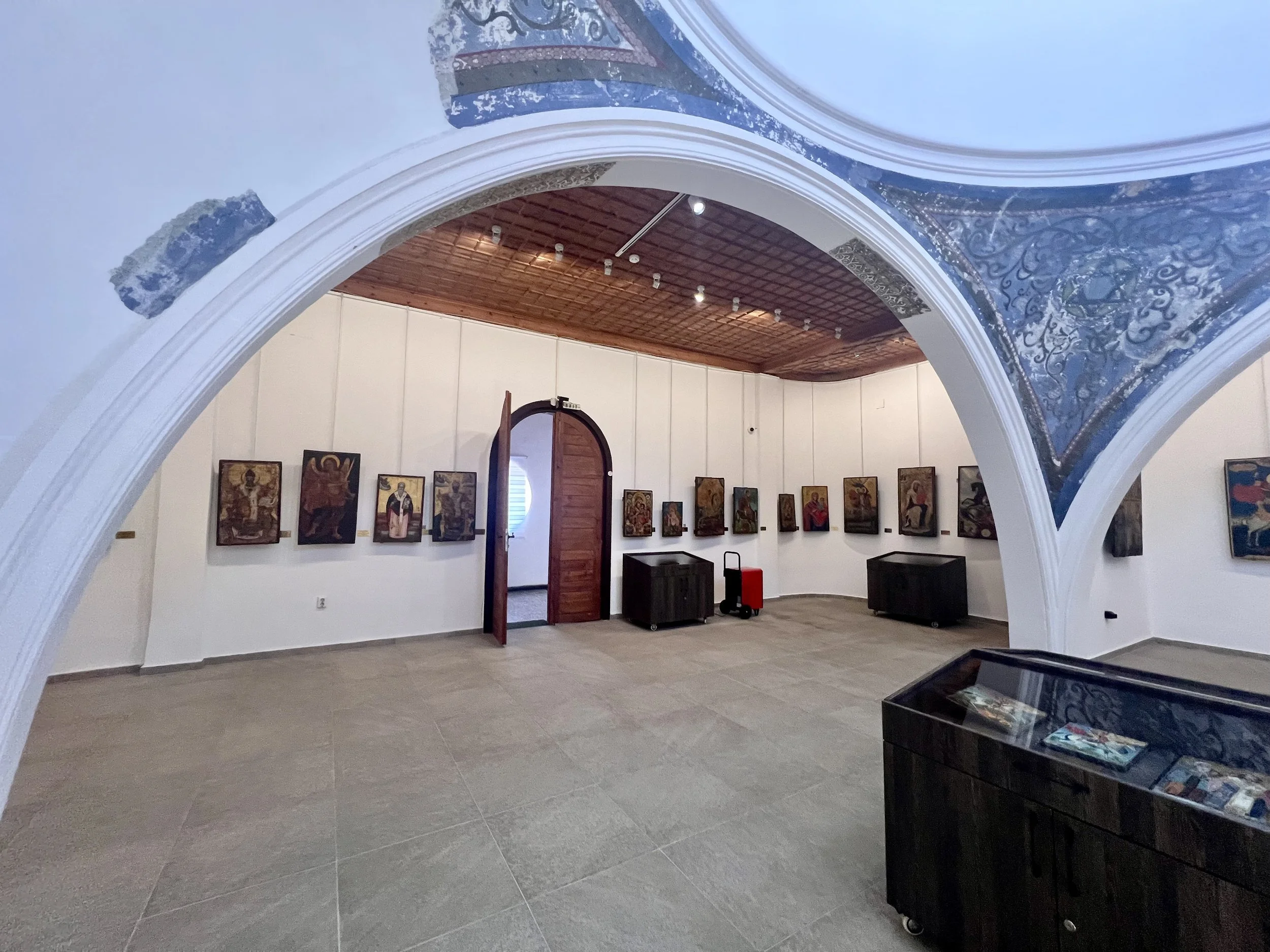 Art gallery with religious icons on white walls and arched ceilings with decorative blue and white floral patterns, wood ceiling, and open door leading to another room.