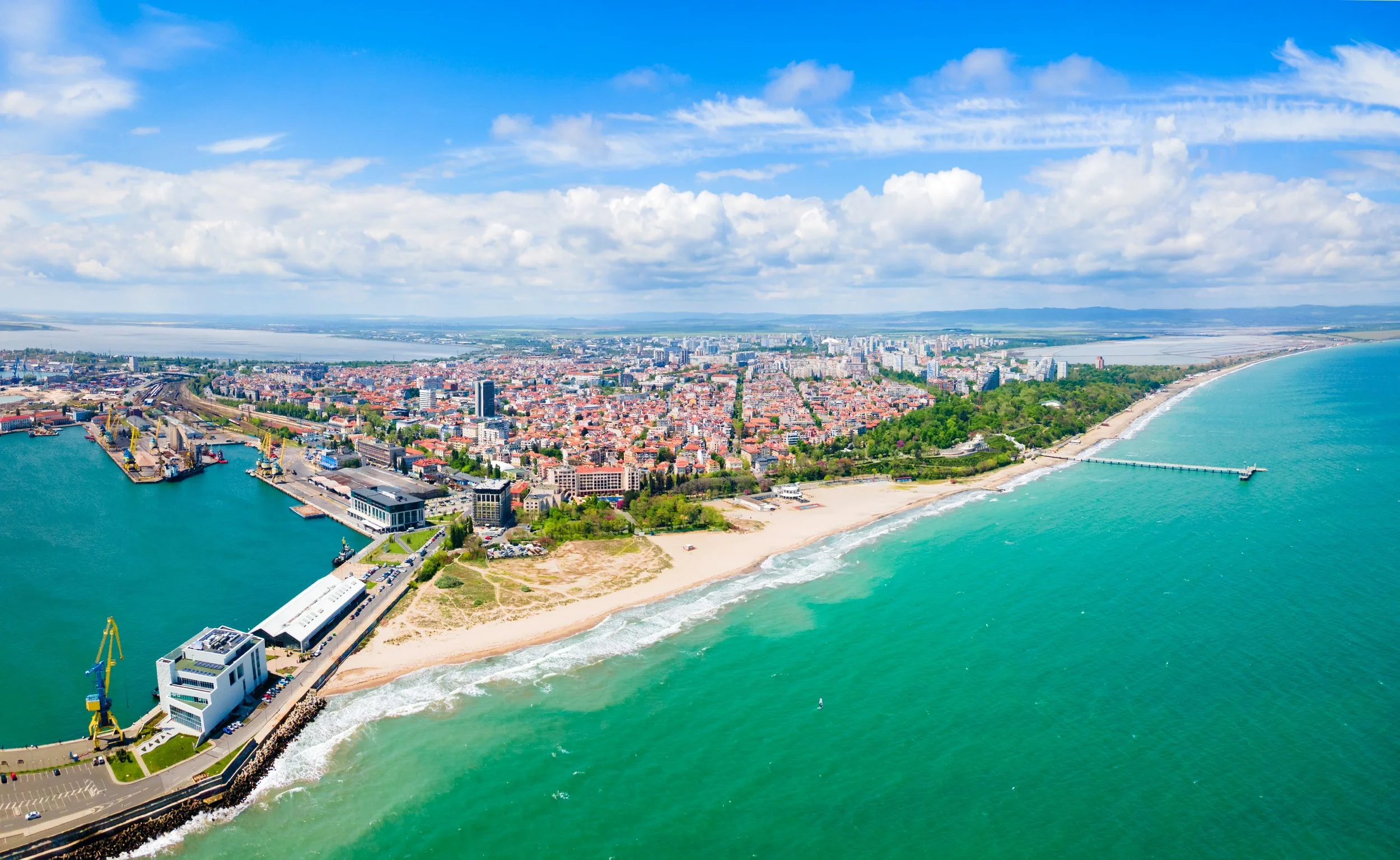 Aerial view of a coastal city with a beach, a pier, and high-rise buildings along the shoreline, with green hills and water in the background under a partly cloudy sky.
