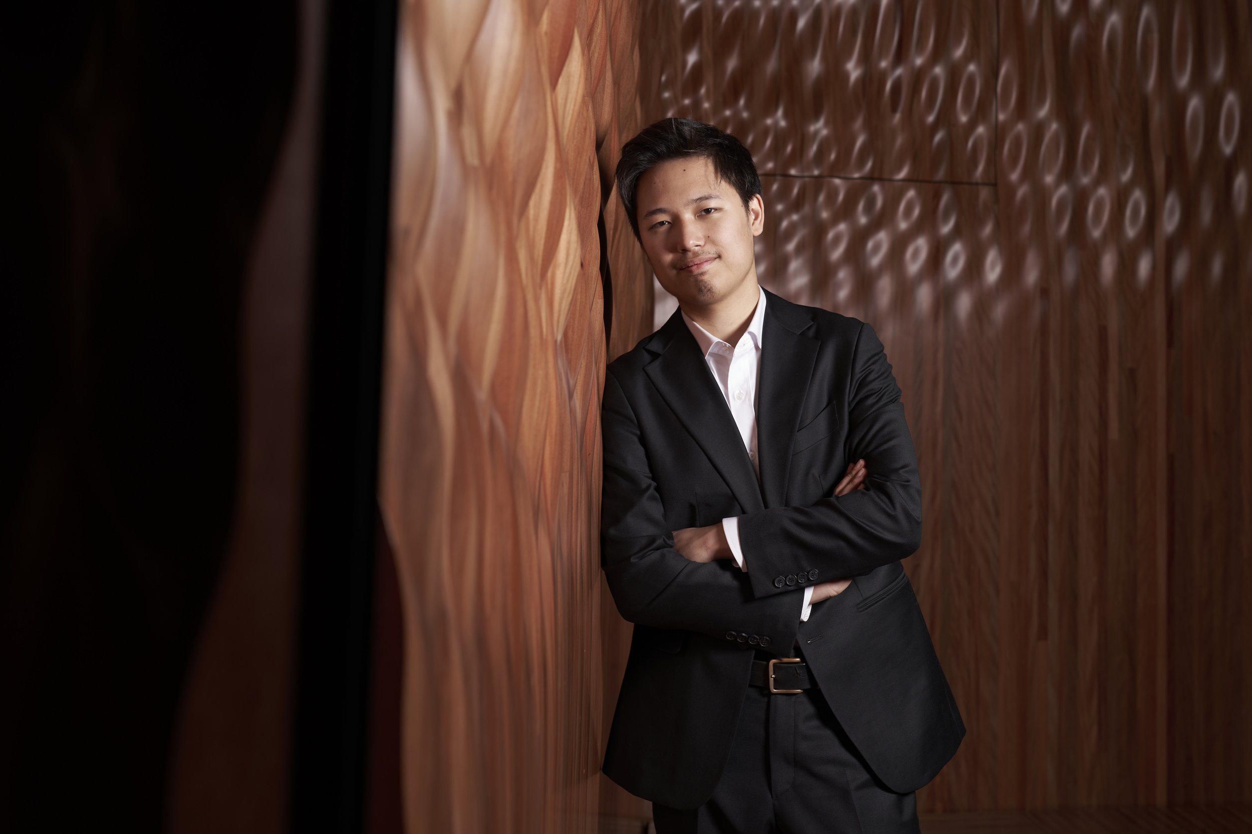 A man in a black suit and white shirt leaning against a wooden wall with crossed arms, looking at the camera.