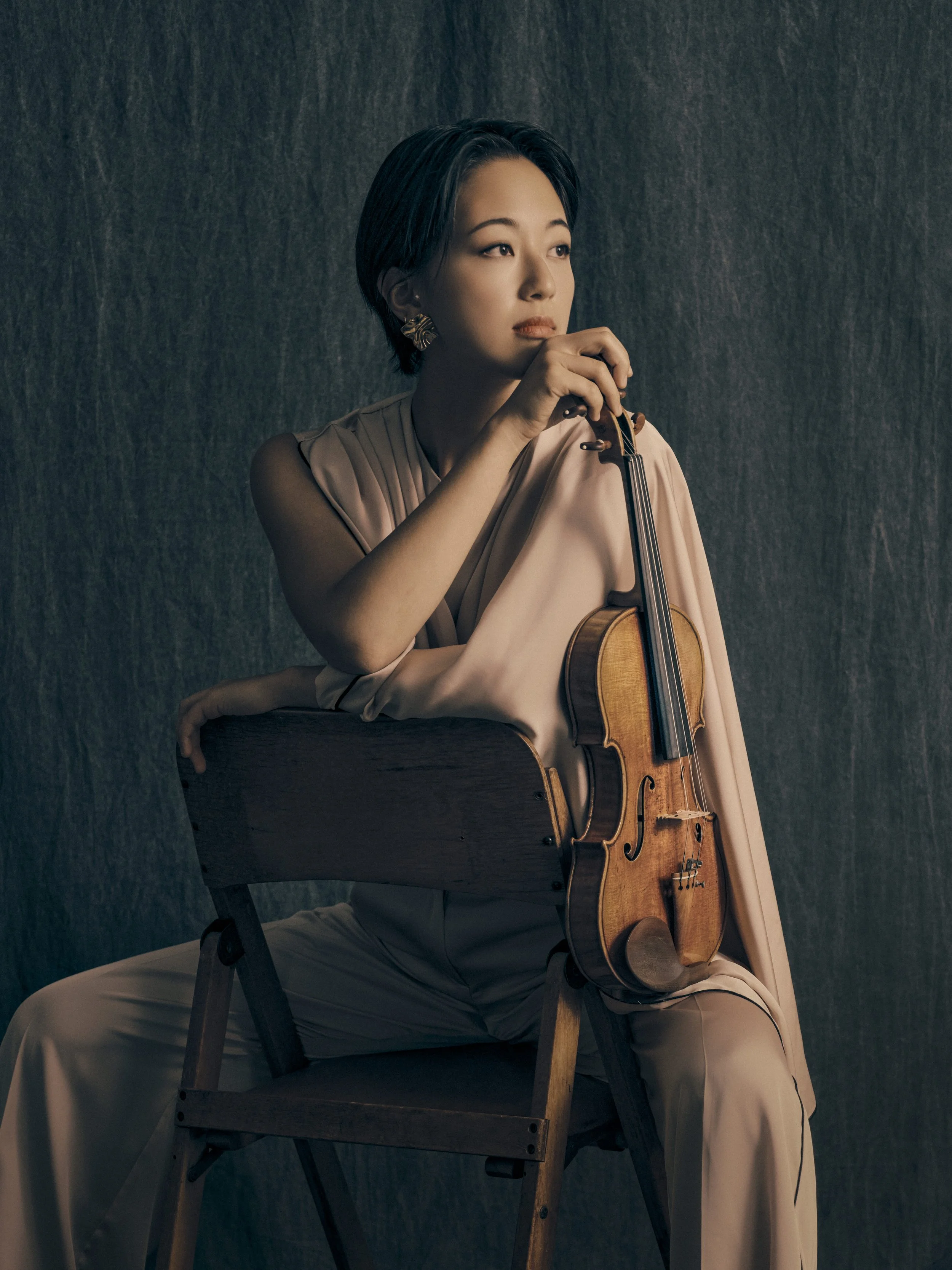 A woman with short black hair and gold earrings sits on a wooden chair, holding a violin and bow, with her gaze directed to the right, against a dark textured background.