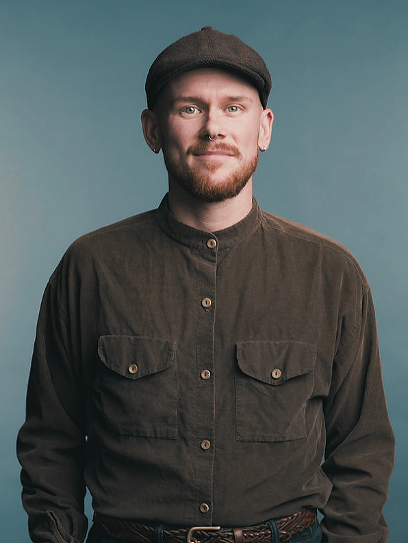 August Waite, counsellor in Victoria, a masculine non-binary person with a red beard standing in a brown shirt and hat in front of a grey background.