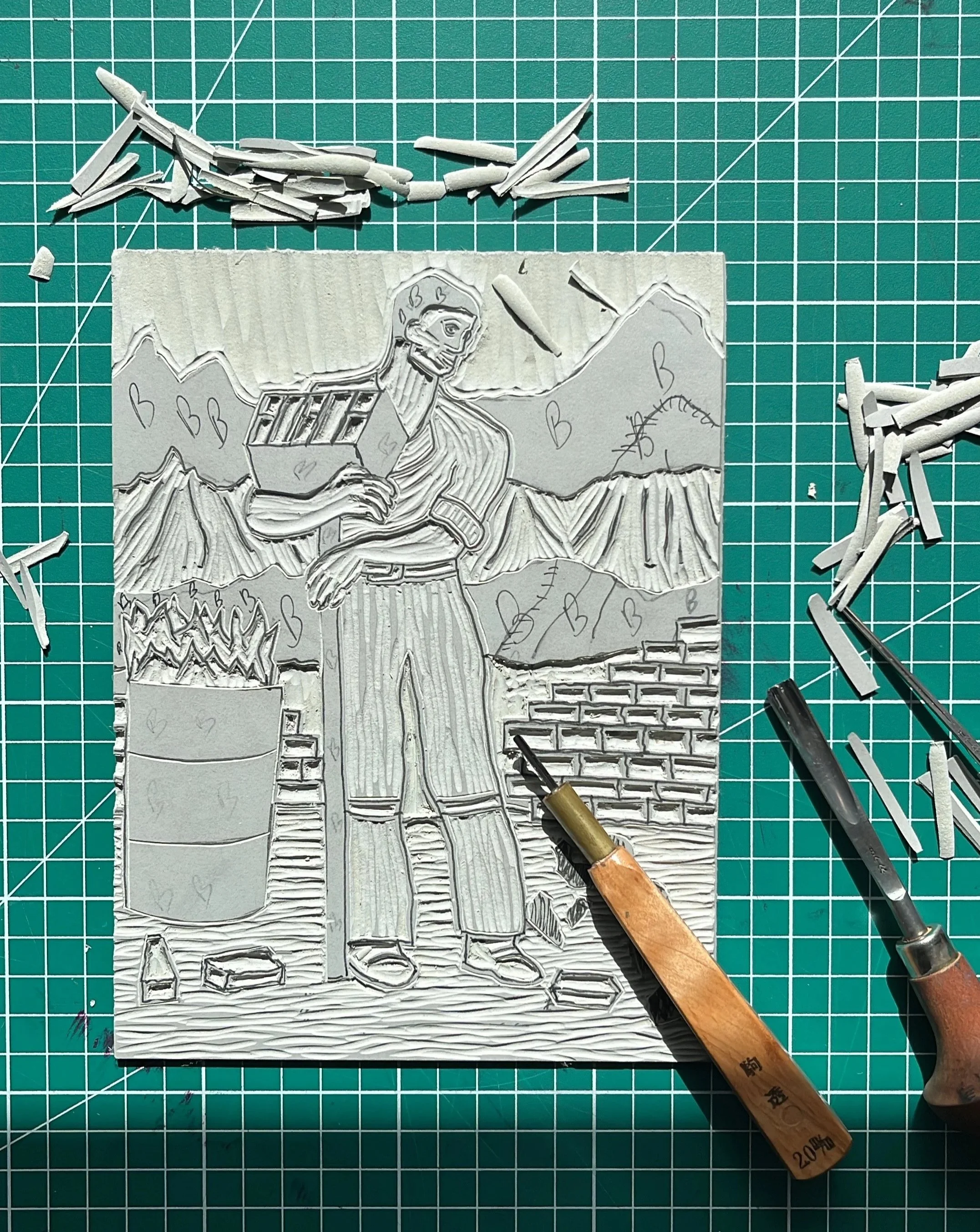 A carved rubber stamp on a green cutting mat with grid lines. The stamp features a hand-drawn depiction of a smiling bird holding a box, with mountain and brick wall illustrations below. Surrounding the stamp are carving tools and pieces of rubber us