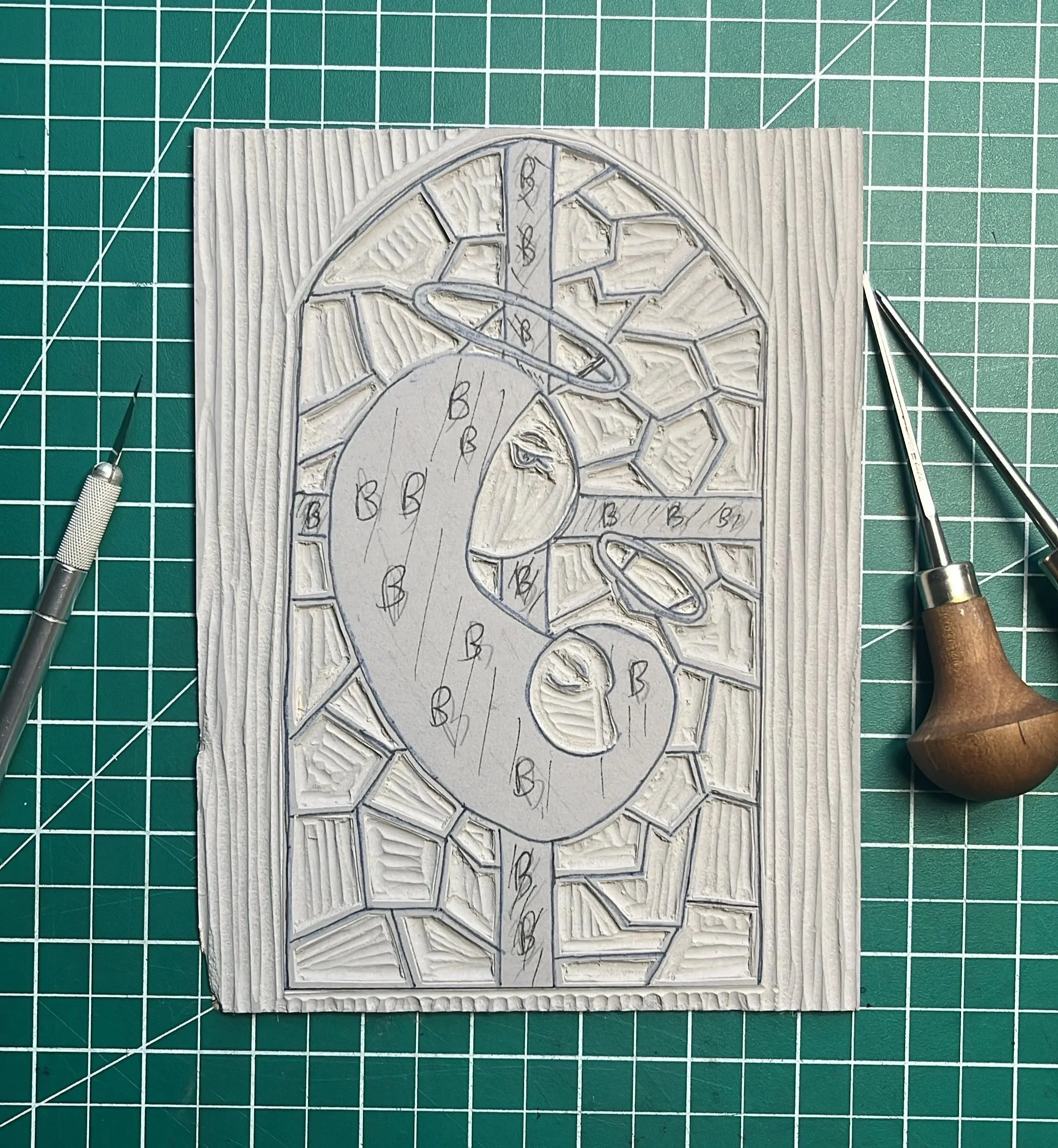 A white textured board with a stained glass window design and handwritten labels. The design includes geometric shapes and labels with letters, numbers, and symbols. There are two precision tools with metallic tips placed on a green cutting mat with 