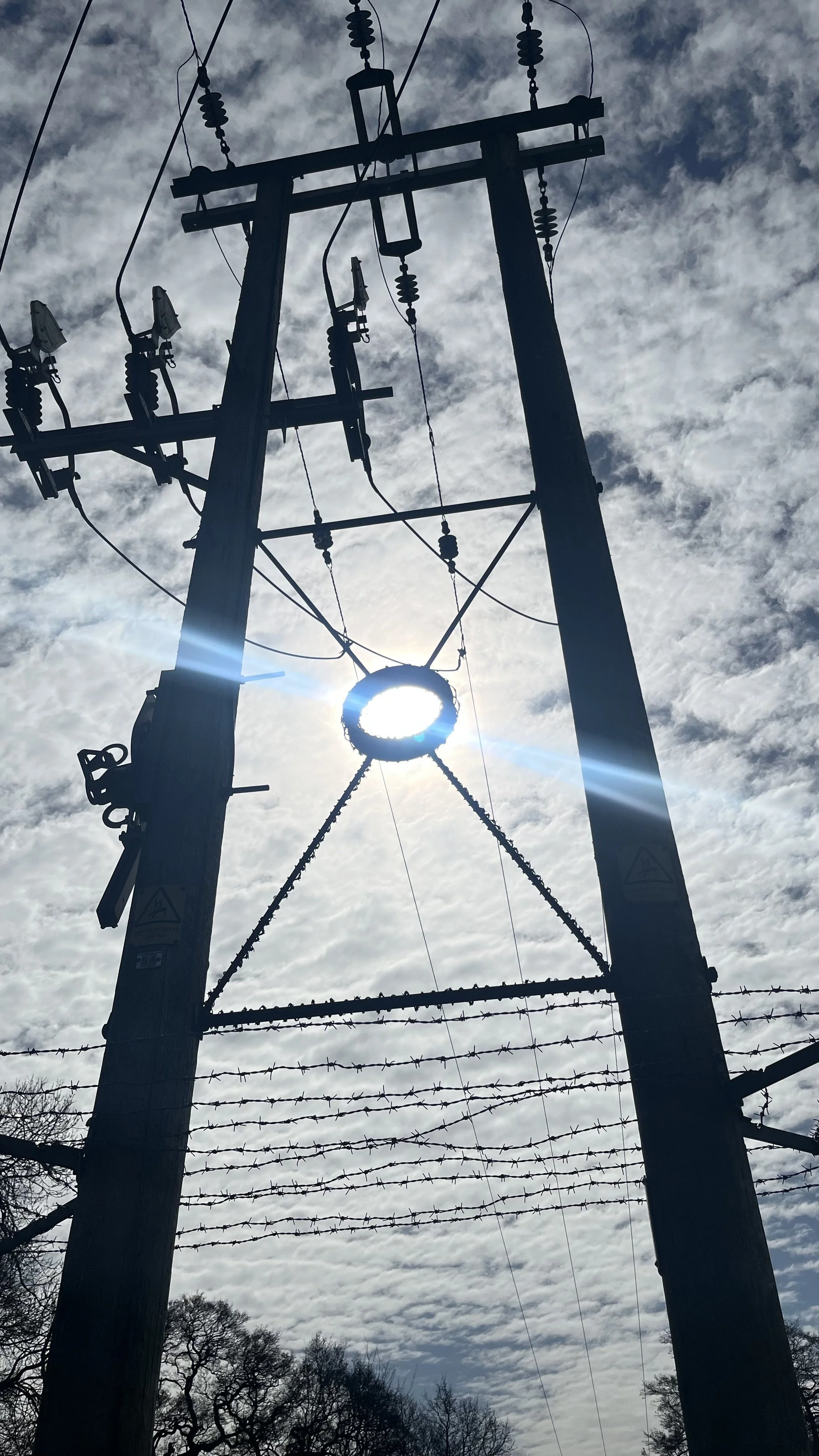 Silhouette of a utility pole with power lines and transformers against a cloudy sky, with the sun partially obscured behind the pole.