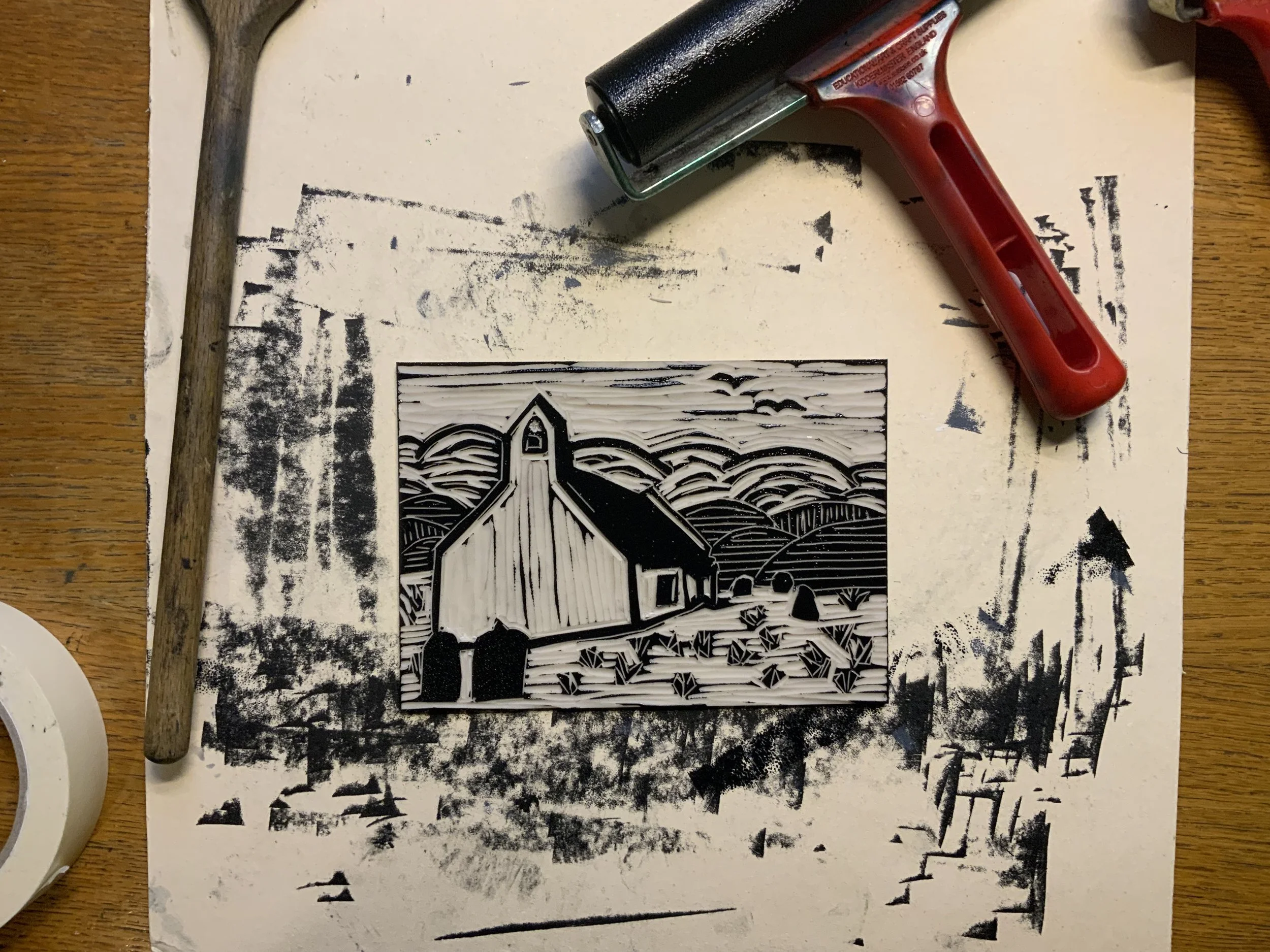 A rubber brayer roller and a linoleum block print of a barn with a field and sky, in black and white, on a work surface with black ink smudges.