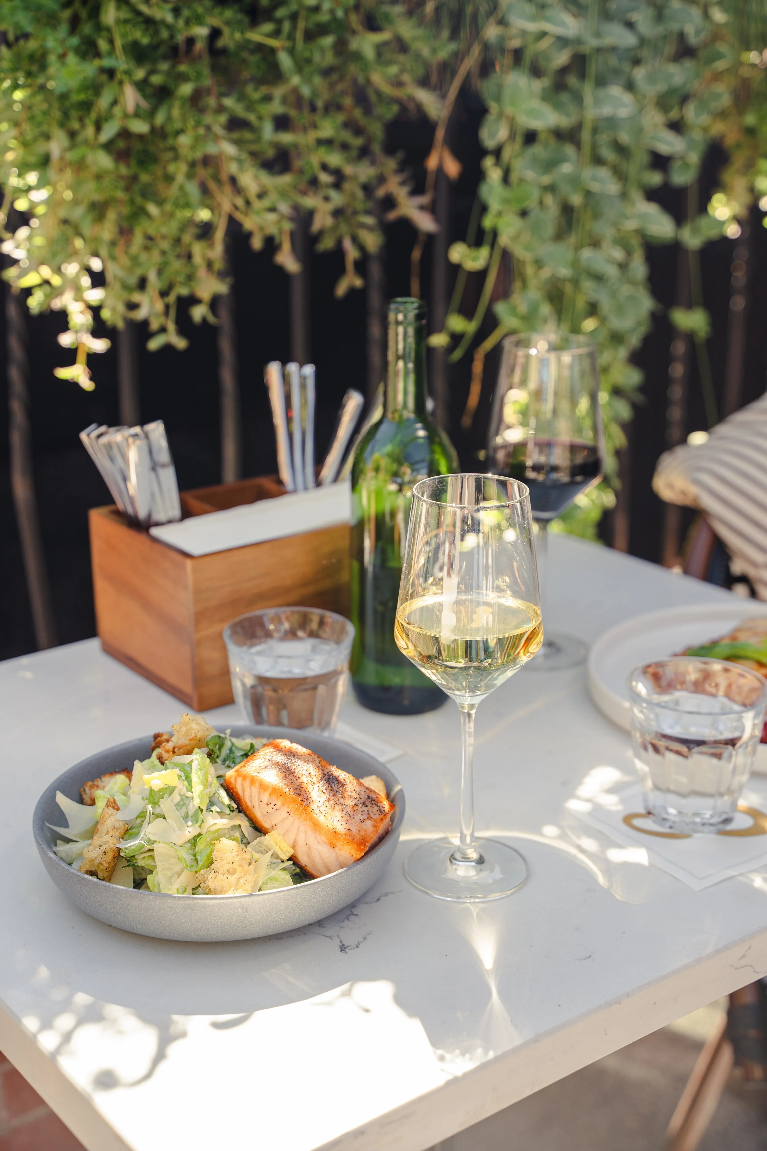 Patio dining in Aspen with a fresh Caesar salad topped with salmon and two glasses of white wine, perfect for a casual lunch in downtown Aspen.