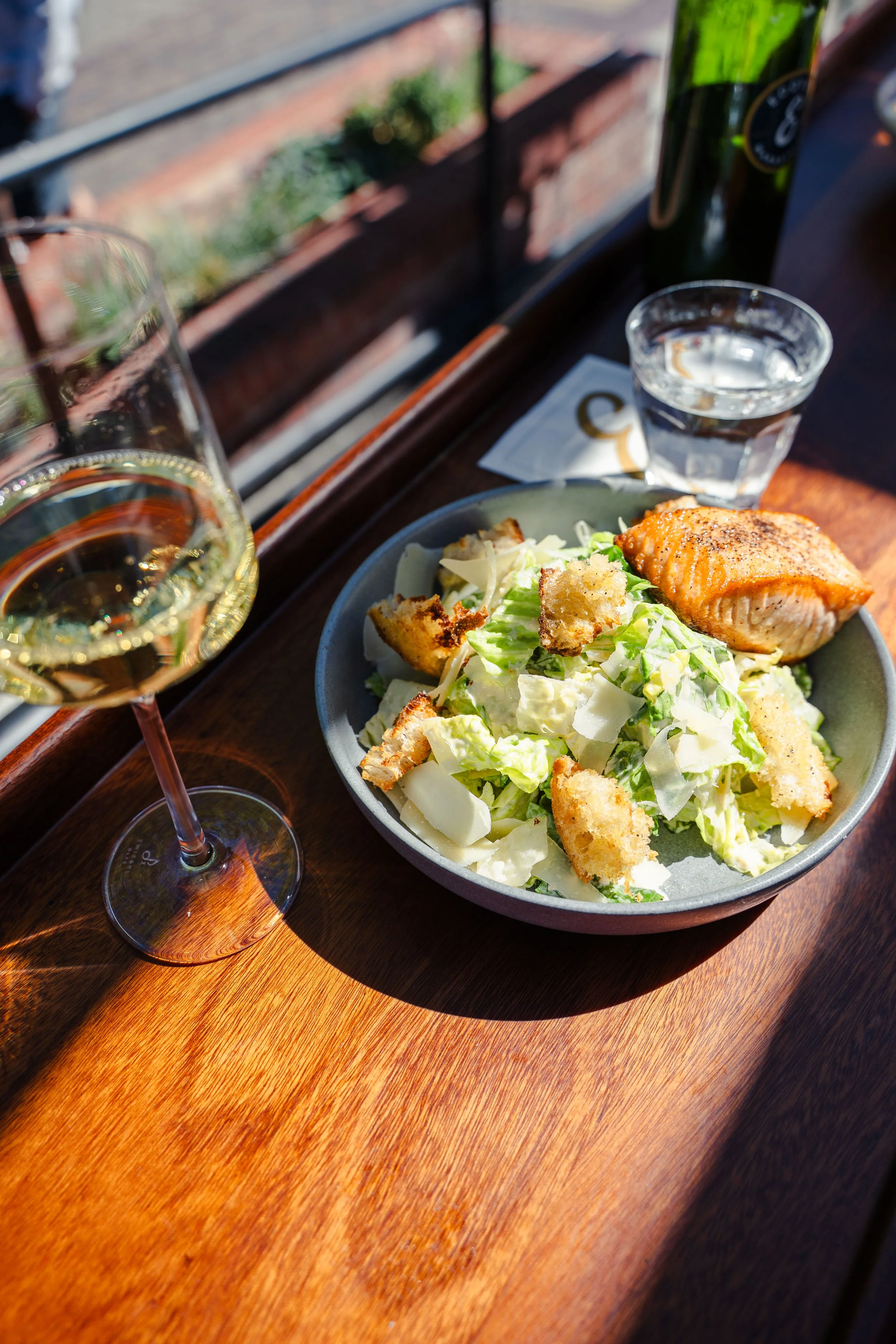 Caesar salad topped with perfectly cooked salmon and paired with a glass of white wine, served at Eddy’s Kitchen’s outdoor bar overlooking the streets of downtown Aspen.