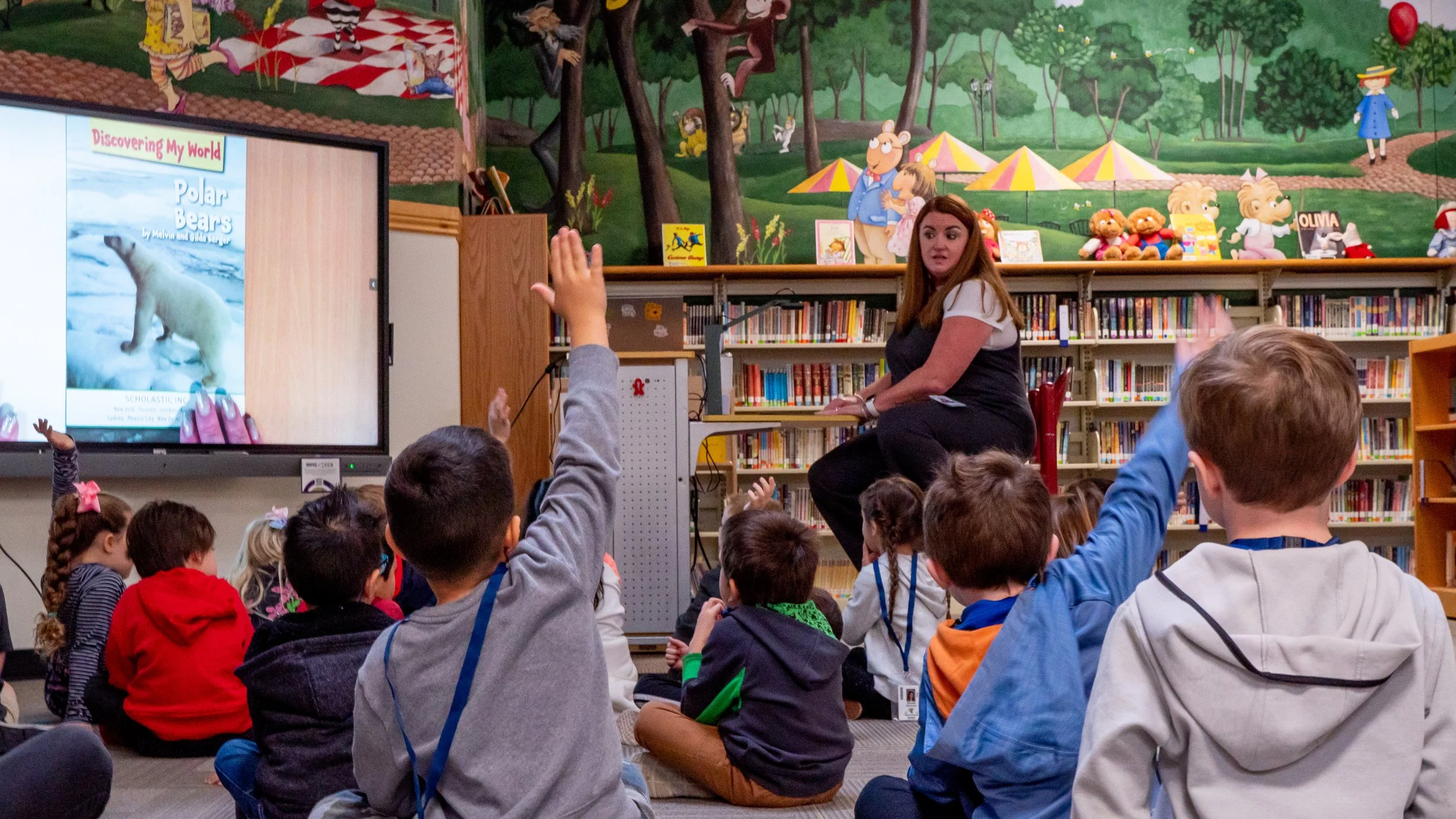 A woman reading to a group of children in a library, with some children raising their hands. The background features colorful murals and shelves of books.