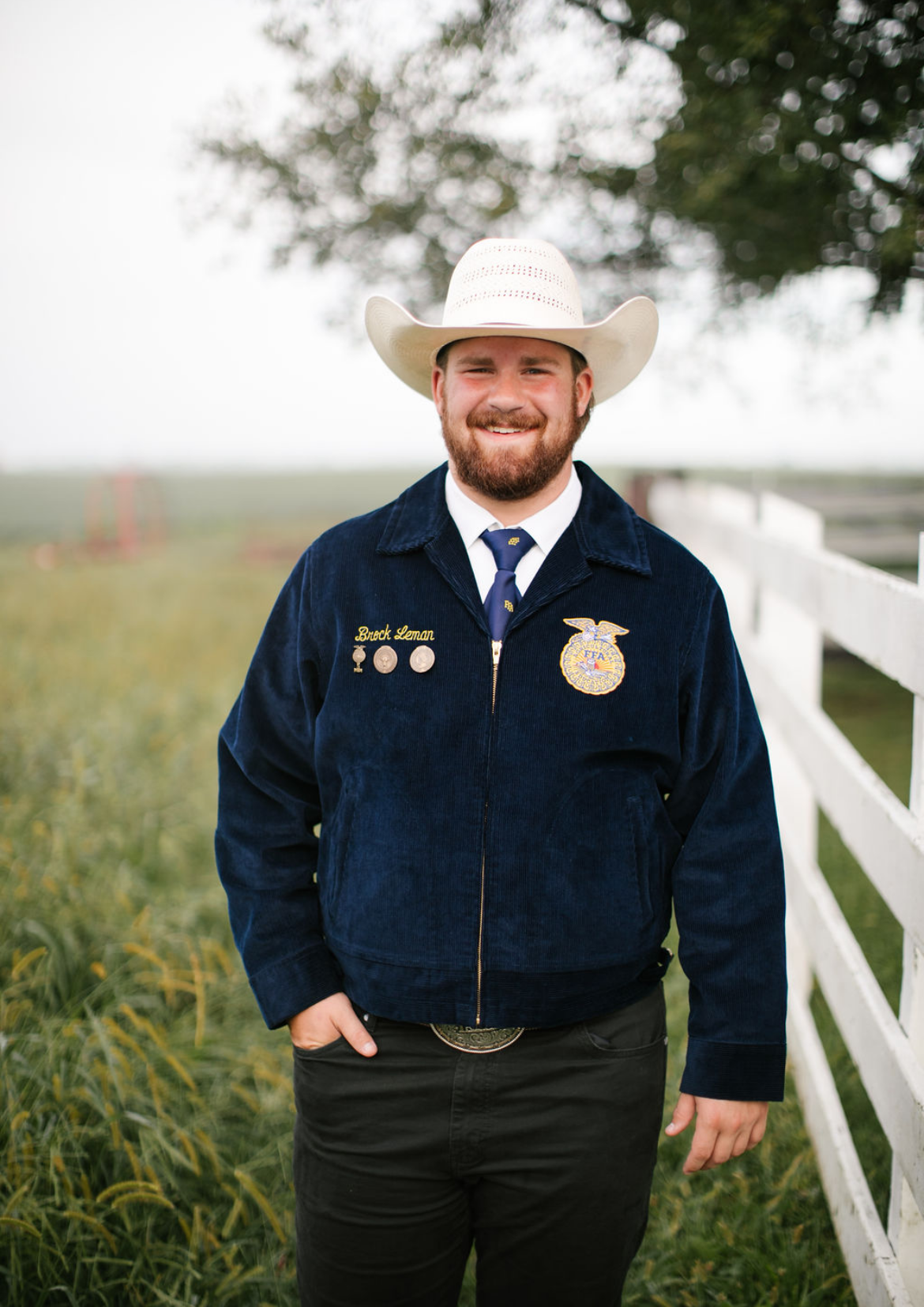A smiling man in a white cowboy hat, navy FFA jacket with medals and badge, standing by a white fence with green field and trees in the background.