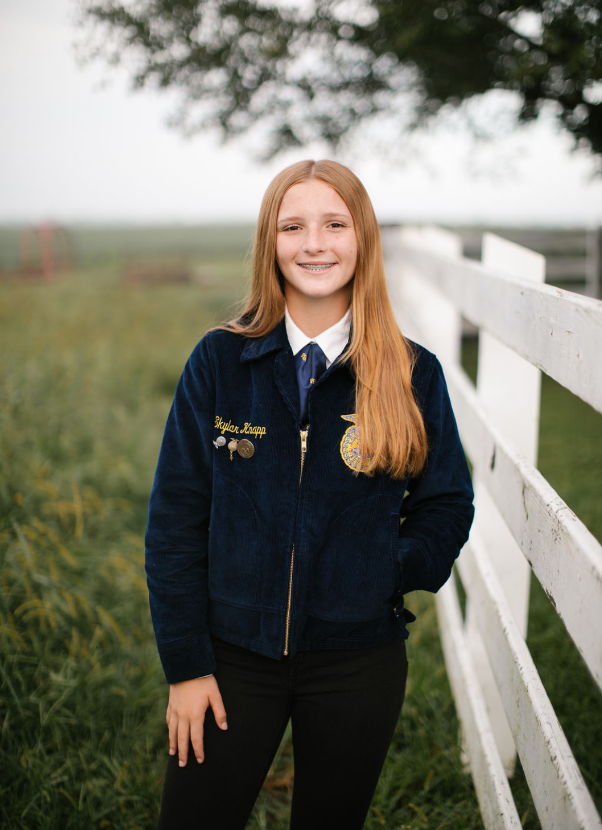 A young girl with long red hair standing outdoors by a white fence, smiling, with a tree and green field in the background.