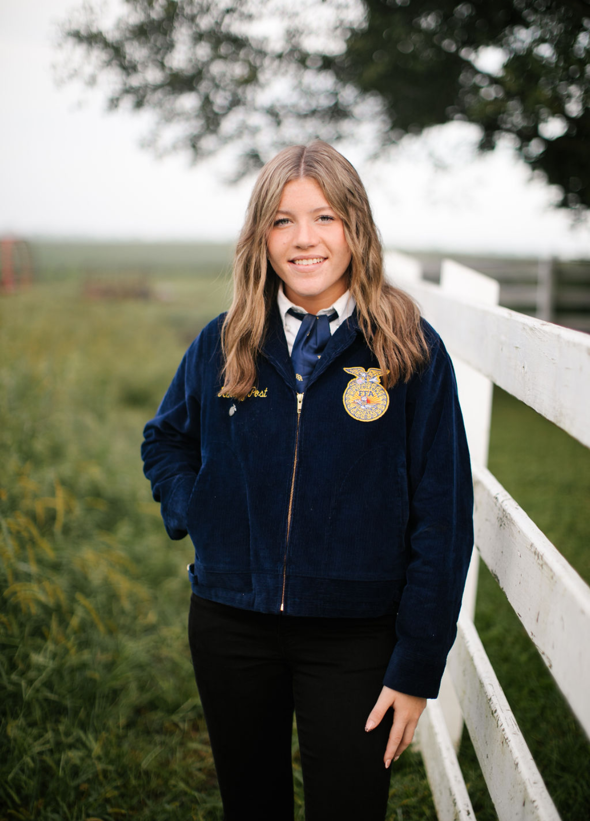 A young woman in an FFA uniform standing outdoors next to a white fence and green grass, smiling at the camera with a blurred background of trees and sky.