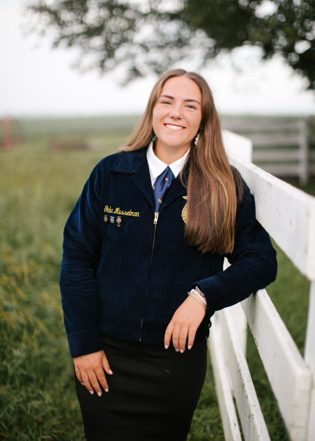 Young woman wearing a FFA jacket, standing outdoors near a white fence, smiling, with green grass and trees in the background.