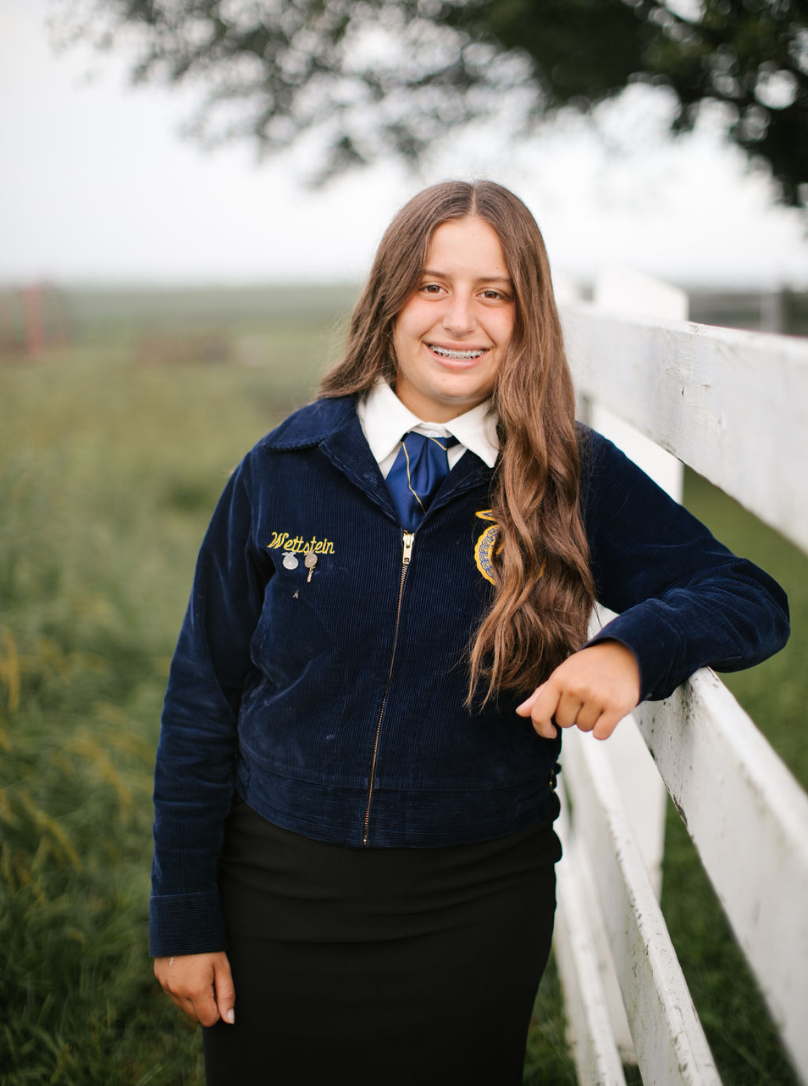 A young girl with long brown hair and braces, wearing a school uniform with a navy blue jacket, white shirt, and navy tie, standing outdoors next to a white wooden fence. She is smiling and resting her arm on the fence with a background of green grass and blurred trees.