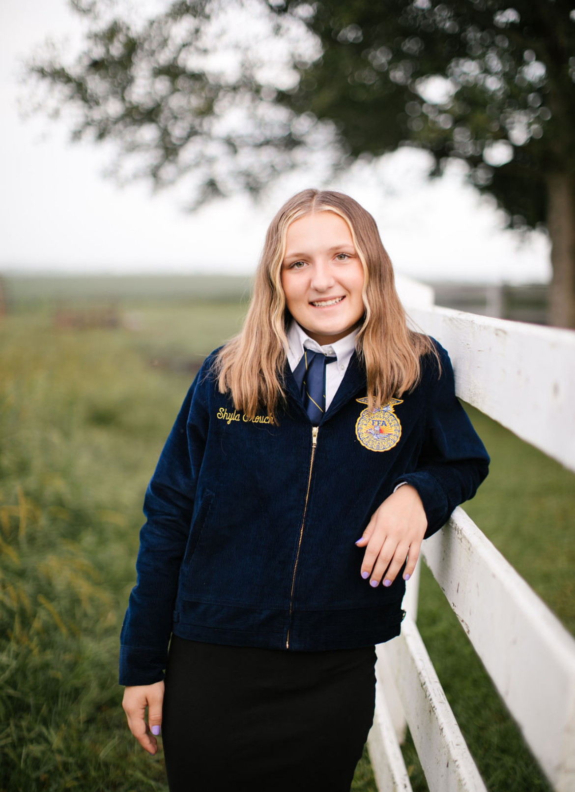 A young girl with blonde hair and a blue FFA jacket standing outdoors by a white fence, smiling, with trees and an open field in the background.