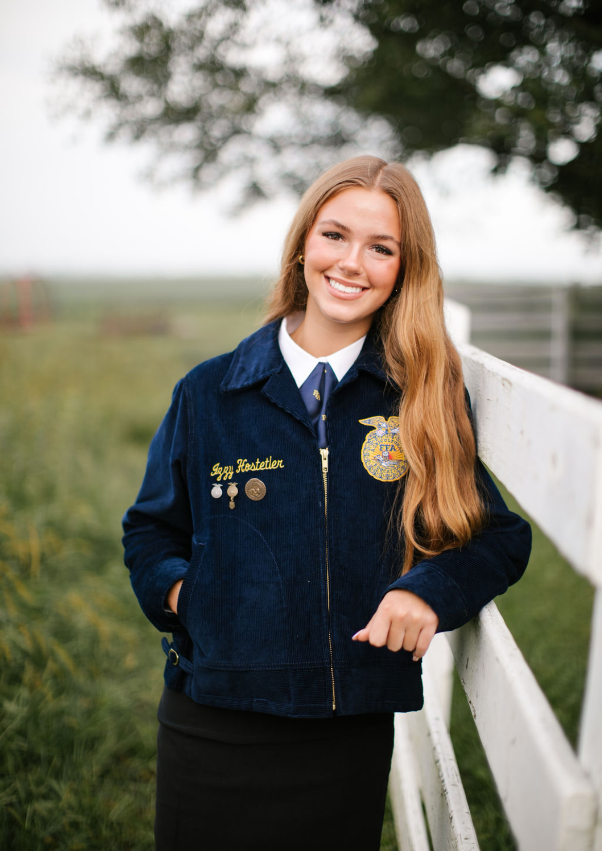 Young woman with long red hair, smiling, wearing a navy blue jacket with FFA emblem and her name, Ann Hestetler, embroidered on it, standing outdoors by a white fence, trees, and green grass.