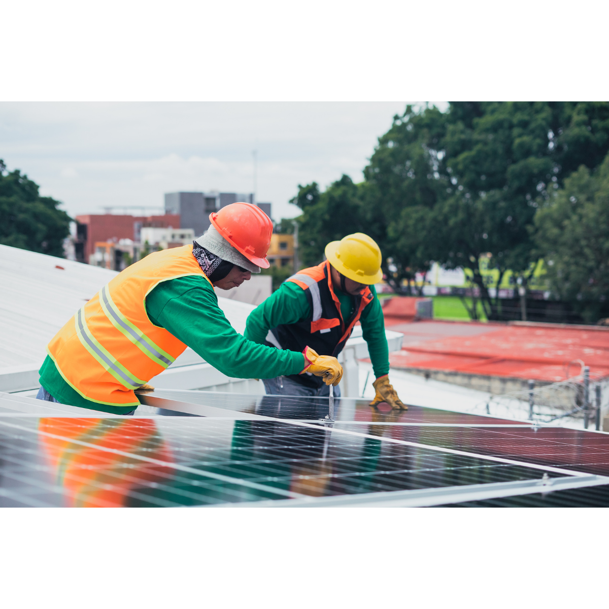 Two workers installing solar panels on a rooftop, wearing safety helmets and vests with a cityscape and trees in the background.