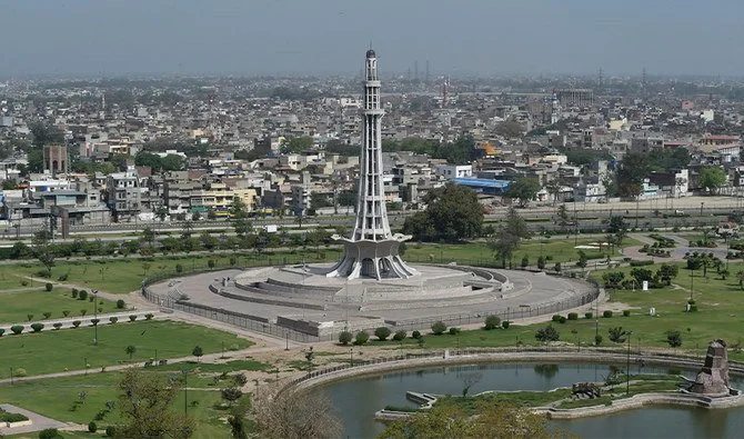 A tall tower with a wide circular base in a large park with a pond, surrounded by city buildings in the background.