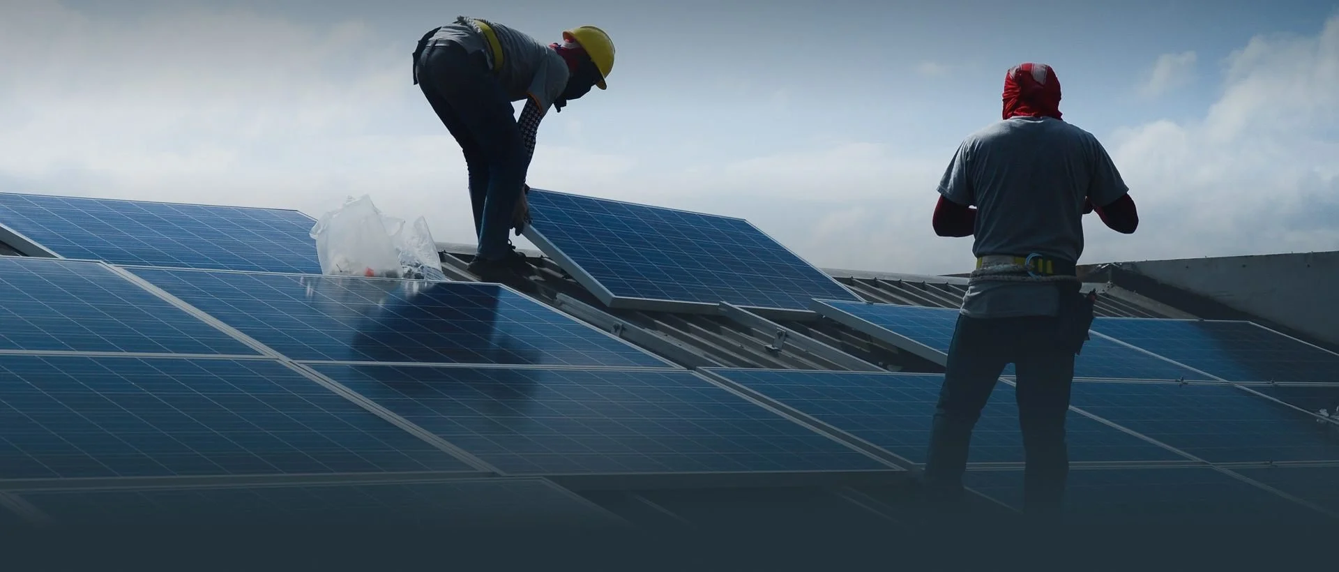 Two workers installing or maintaining solar panels on a roof, with one person wearing a yellow safety helmet and the other with a red cloth on their head, under a partly cloudy sky.