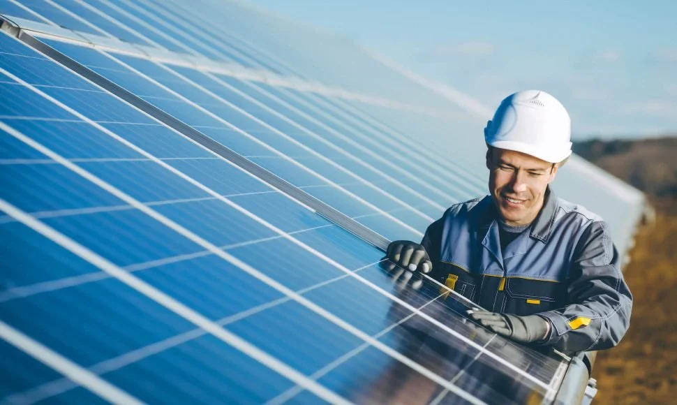 A worker in a white safety helmet and gray work uniform installs or maintains solar panels outdoors on a sunny day.