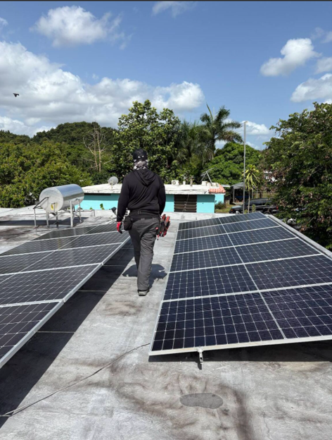 Person walking on a rooftop with solar panels, holding tools, with trees and a blue sky in the background.