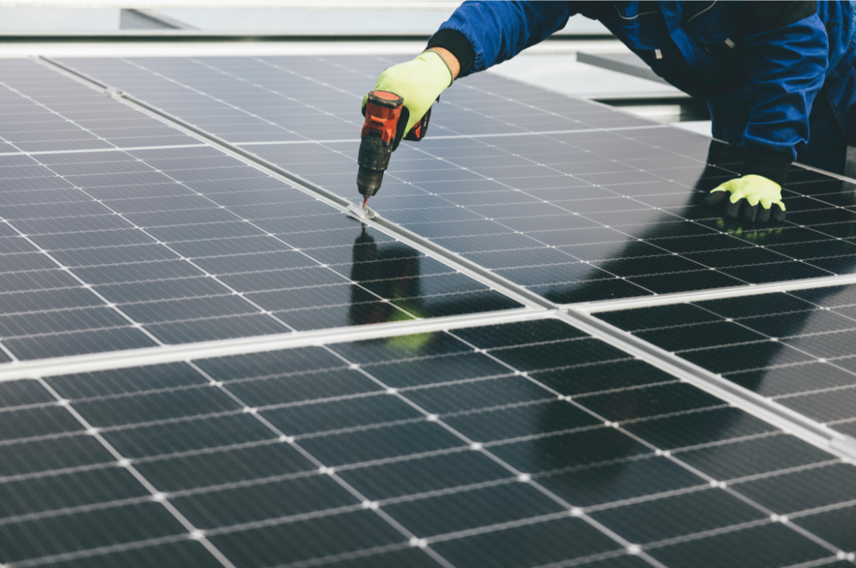 A worker installing or repairing solar panels on a rooftop, using a cordless drill, wearing gloves and a blue jacket.