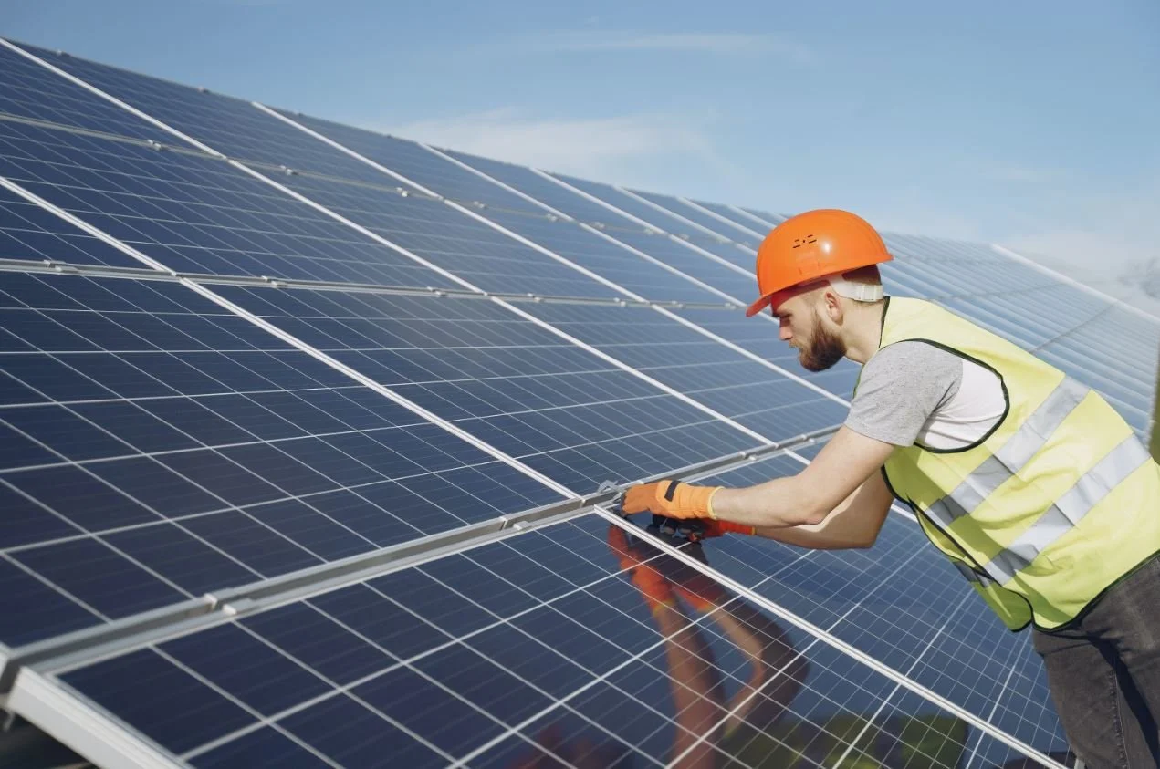 A man wearing a safety helmet and vest working on solar panels outdoors on a sunny day.