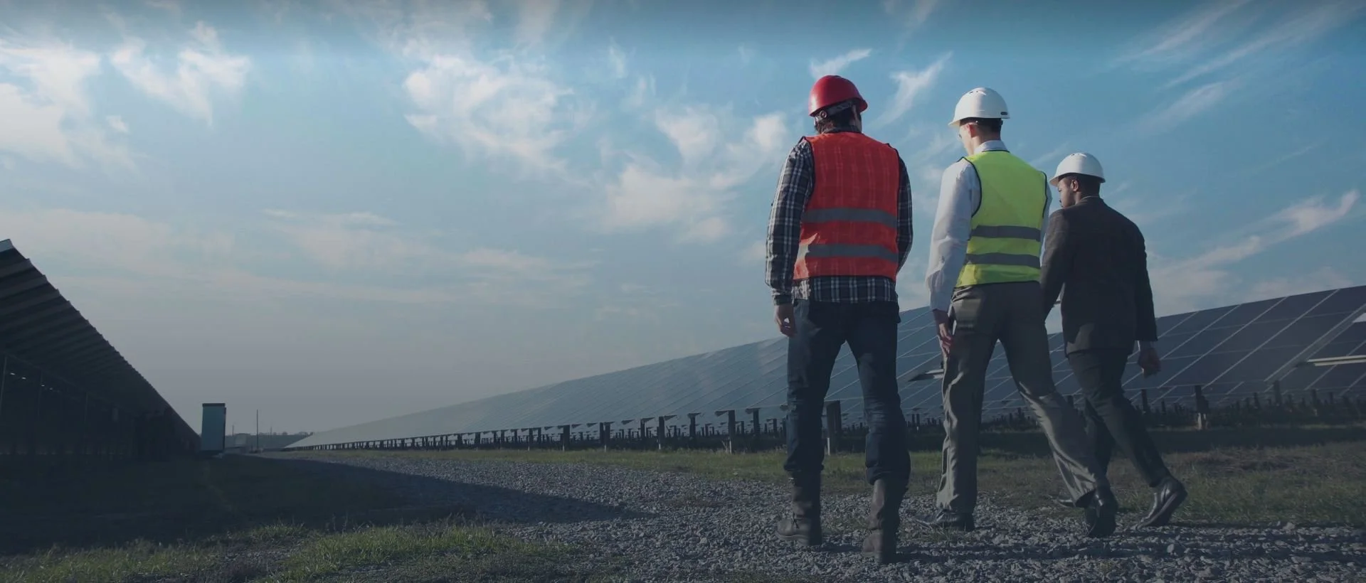 Three people in safety vests and helmets walking through a solar farm with solar panels under a partly cloudy sky.