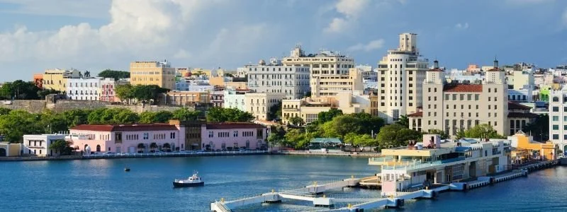 City skyline with buildings along a river, including a pink historic fort and a ferry dock.