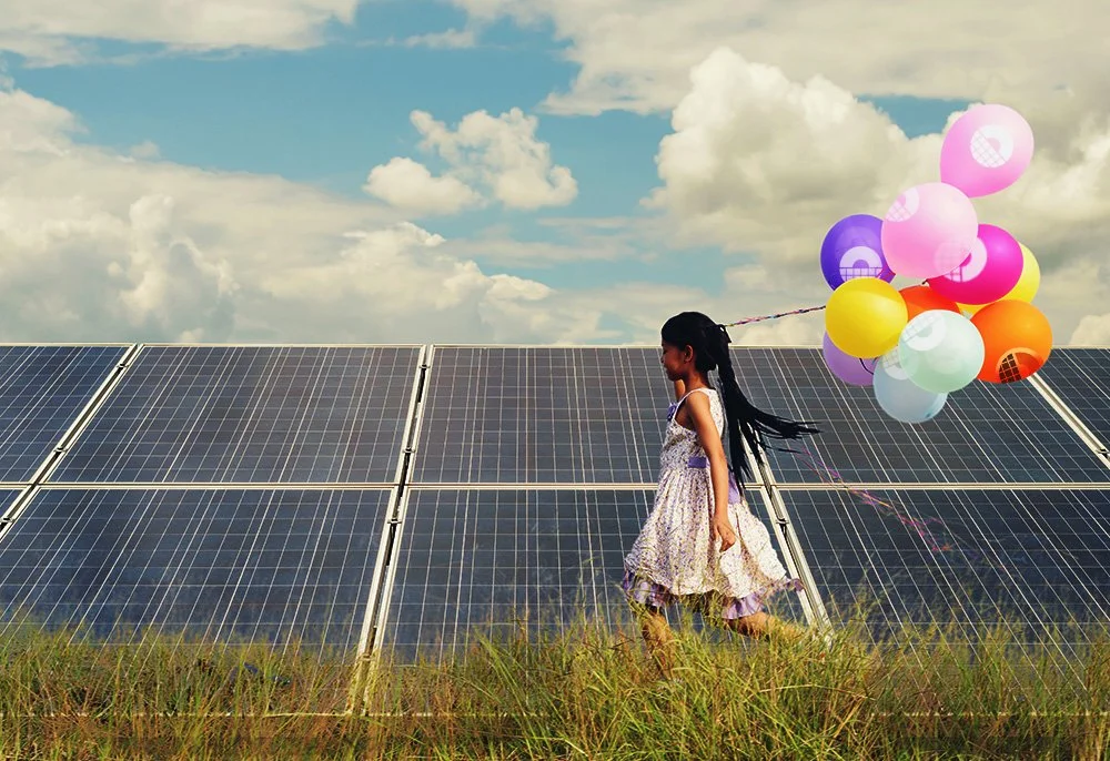 A young girl in a floral dress walking in a grassy field beside solar panels, holding colorful balloons against a partly cloudy sky.