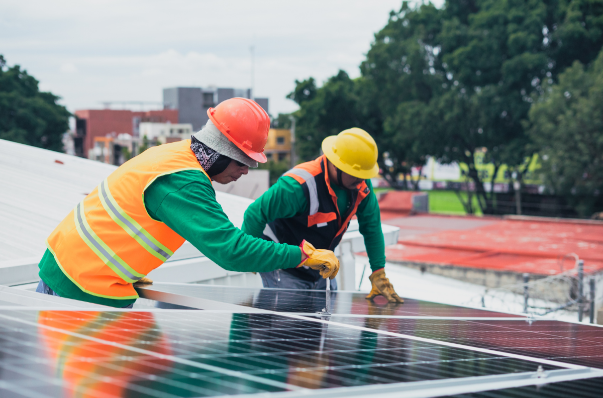 Two workers in safety helmets and reflective vests installing solar panels on a roof.