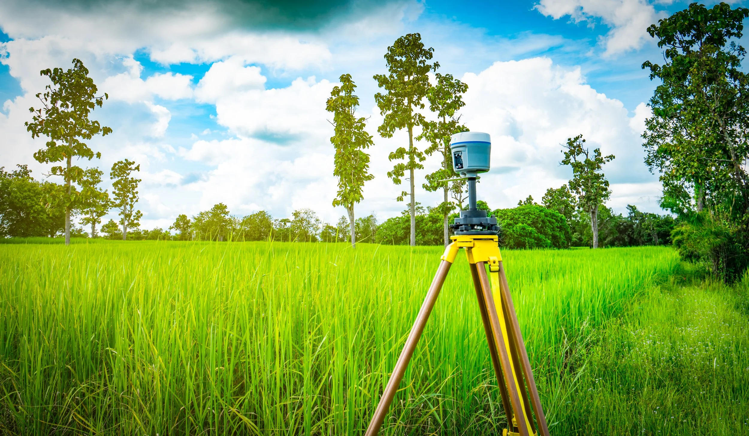 A surveying instrument mounted on a tripod in a grassy field with trees and a partly cloudy sky in the background.