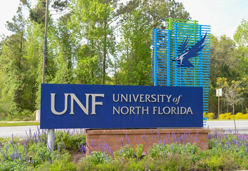 Sign for the University of North Florida with a blue logo and a decorative blue and green structure behind it, surrounded by purple flowers and green trees.