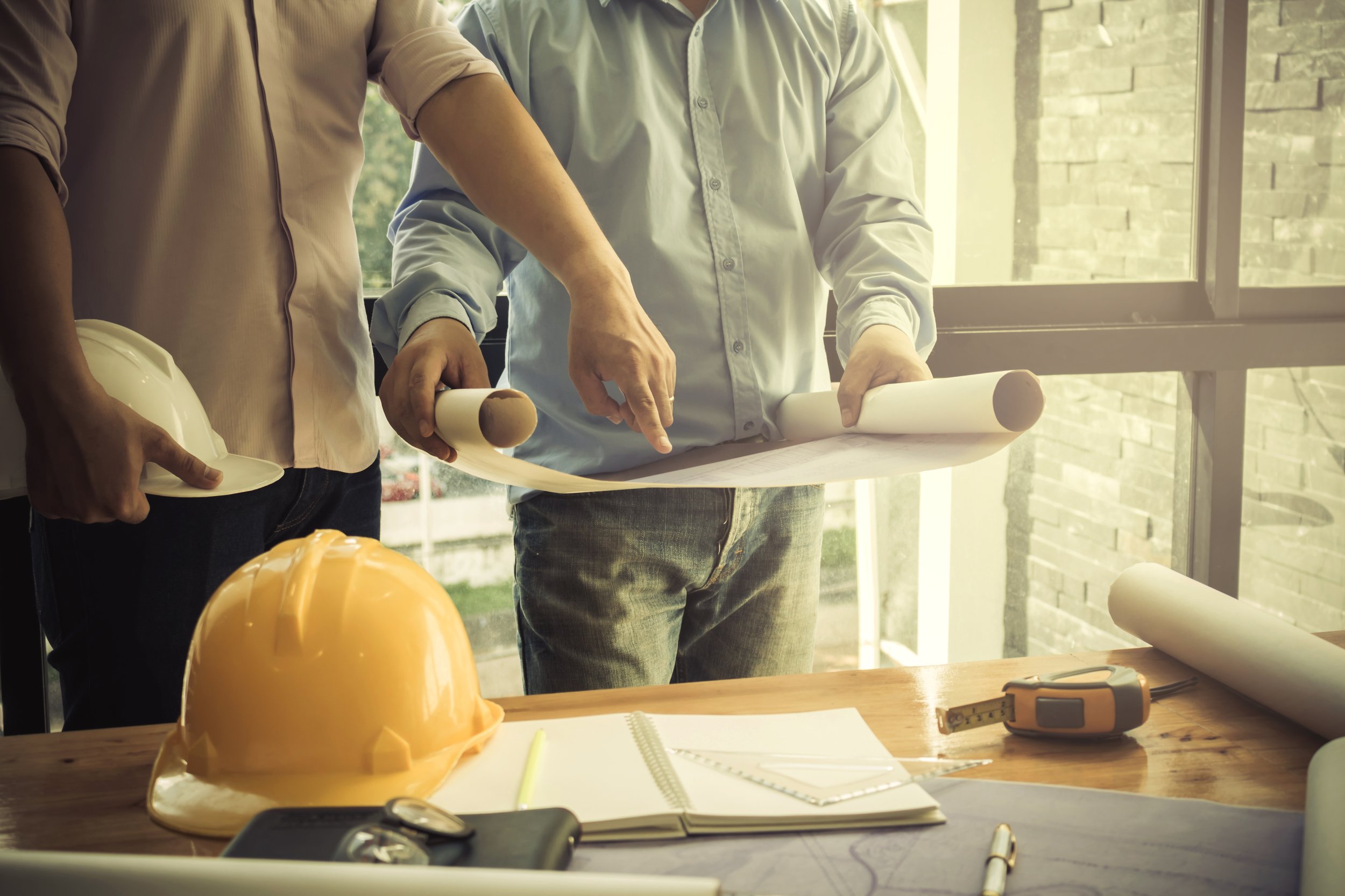 Two men are examining architectural blueprints on a table while standing near a window. One man is pointing at the blueprints. Construction hats, a measuring tape, and notebooks are on the table.