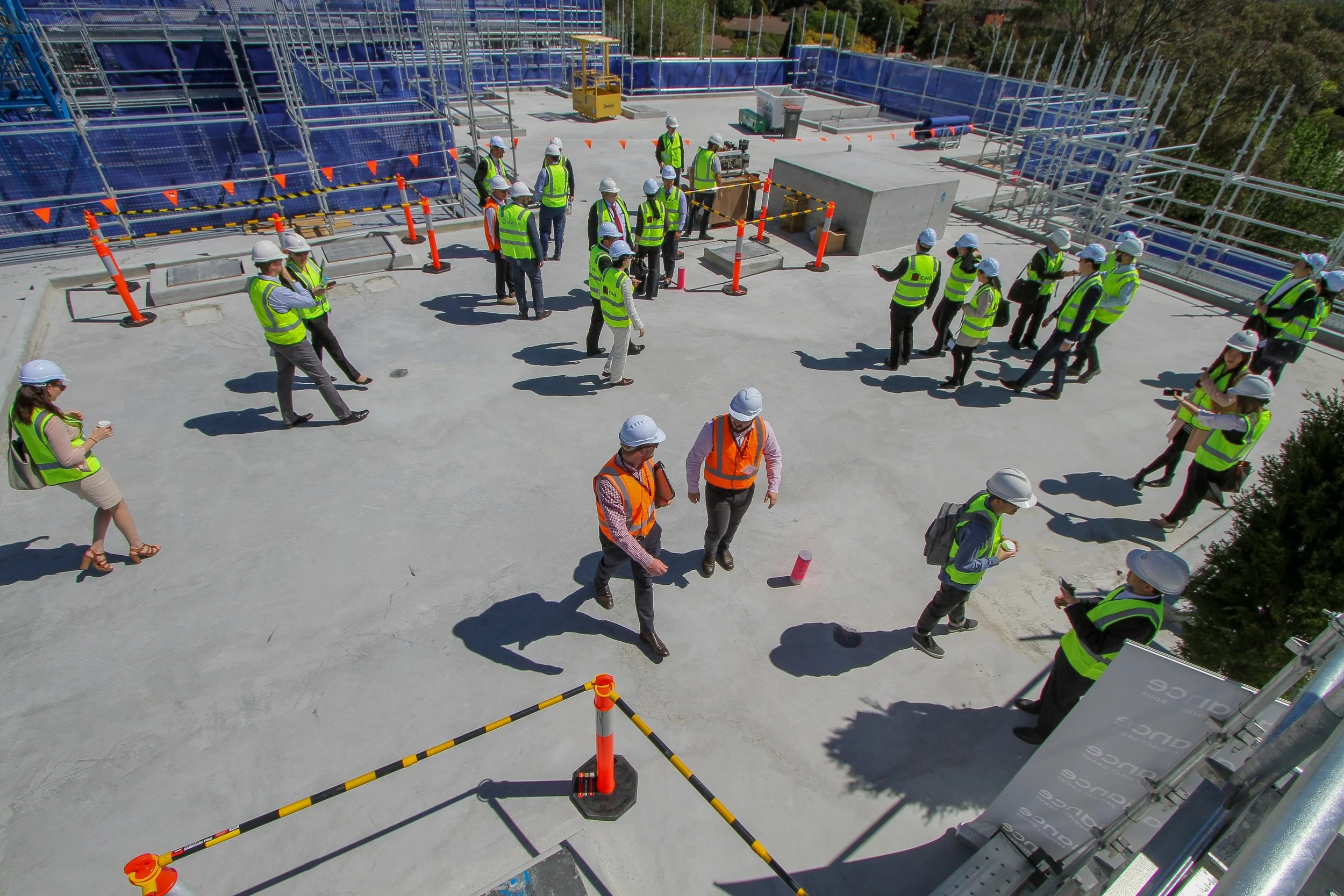 Construction site with multiple workers wearing safety vests and helmets, standing on a concrete area surrounded by safety barriers, with scaffolding and construction materials in the background.