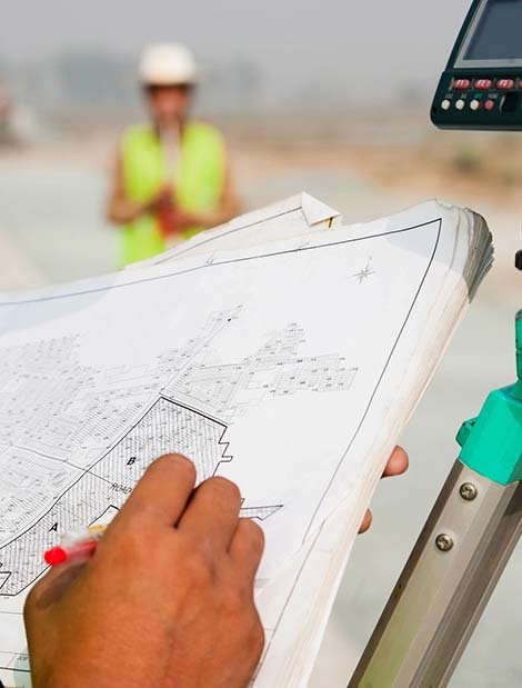A person holding a blueprint with a red pen, with construction equipment in the background, and a person wearing a hard hat and safety vest in the distance.