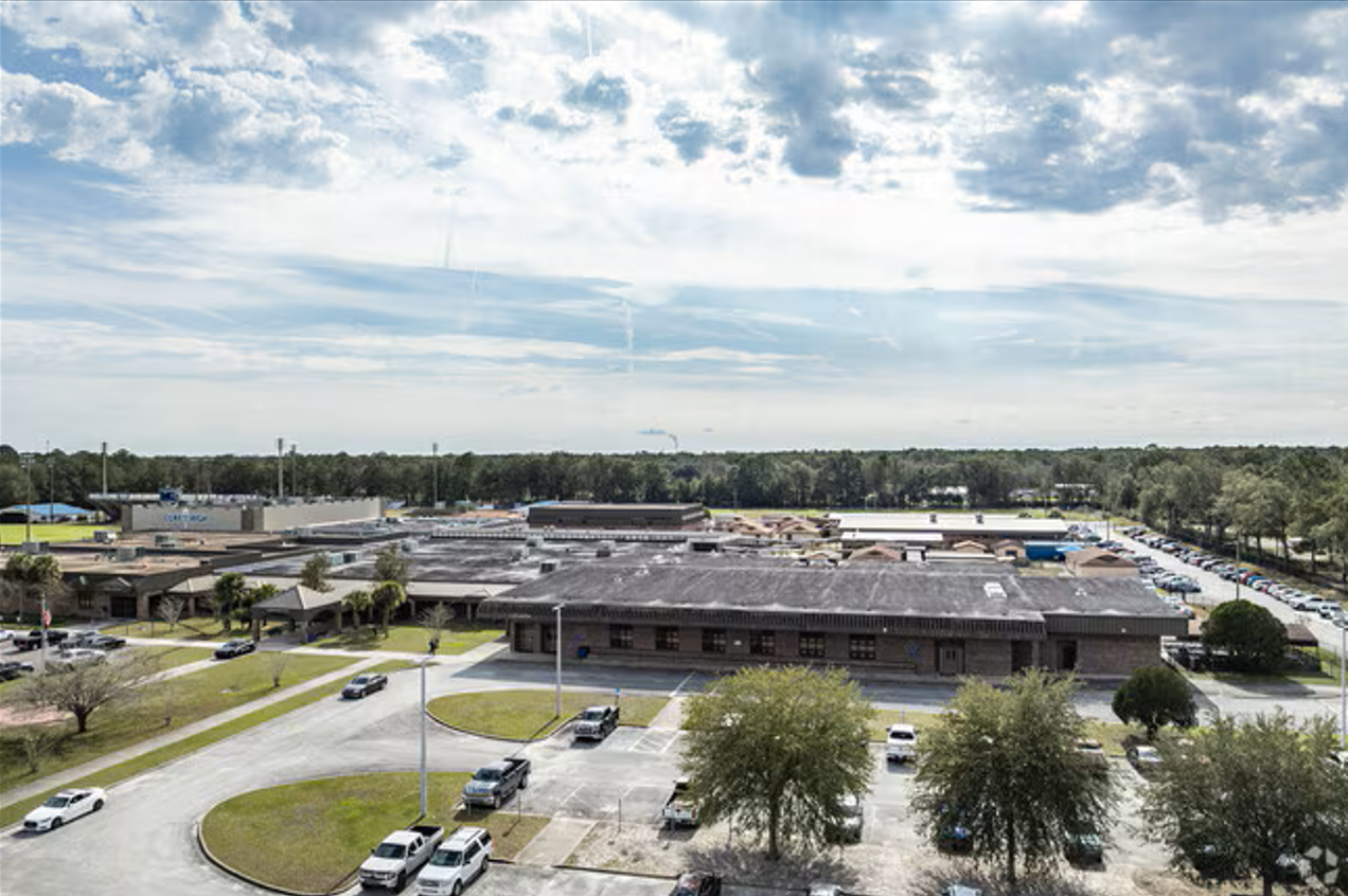 A large single-story shopping center with a parking lot and several trees in the foreground, under a partly cloudy sky.