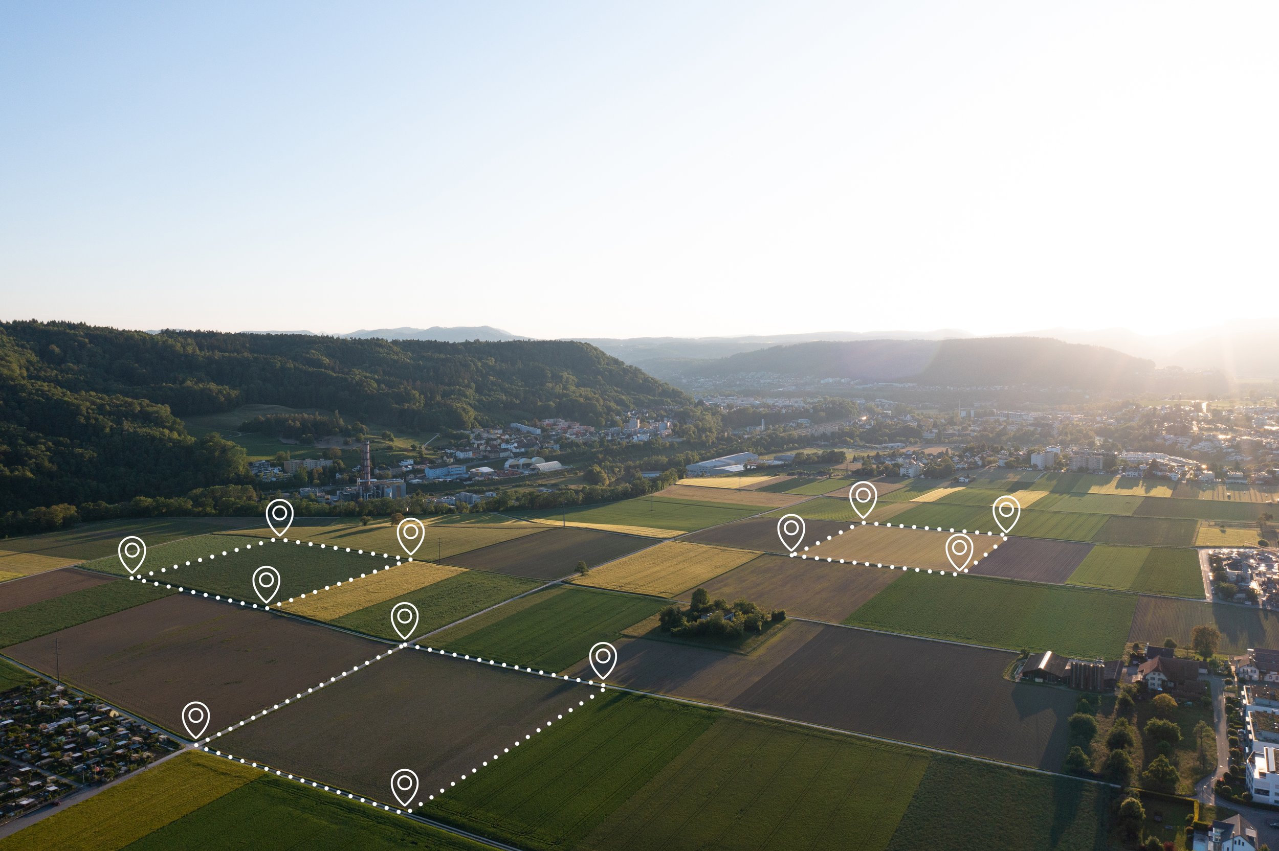Aerial view of a rural landscape with agricultural fields, small town buildings, and green hills, marked with location pins connected by dotted lines, during sunset.