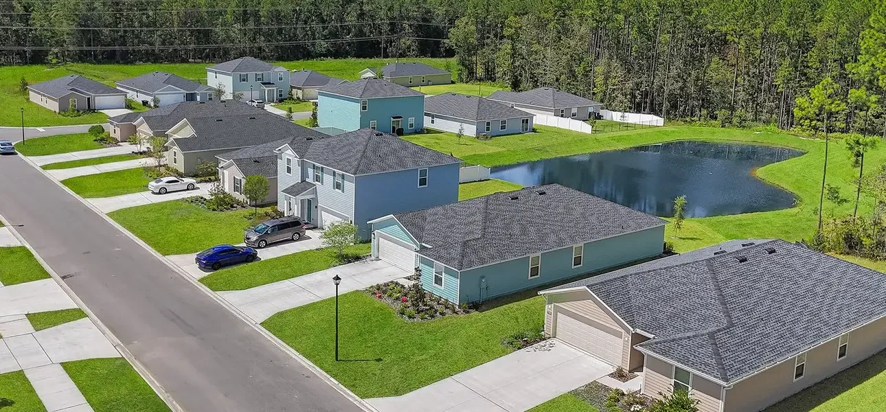 Aerial view of a suburban neighborhood with multiple houses, green lawns, parked cars, a small lake, and trees in the background.