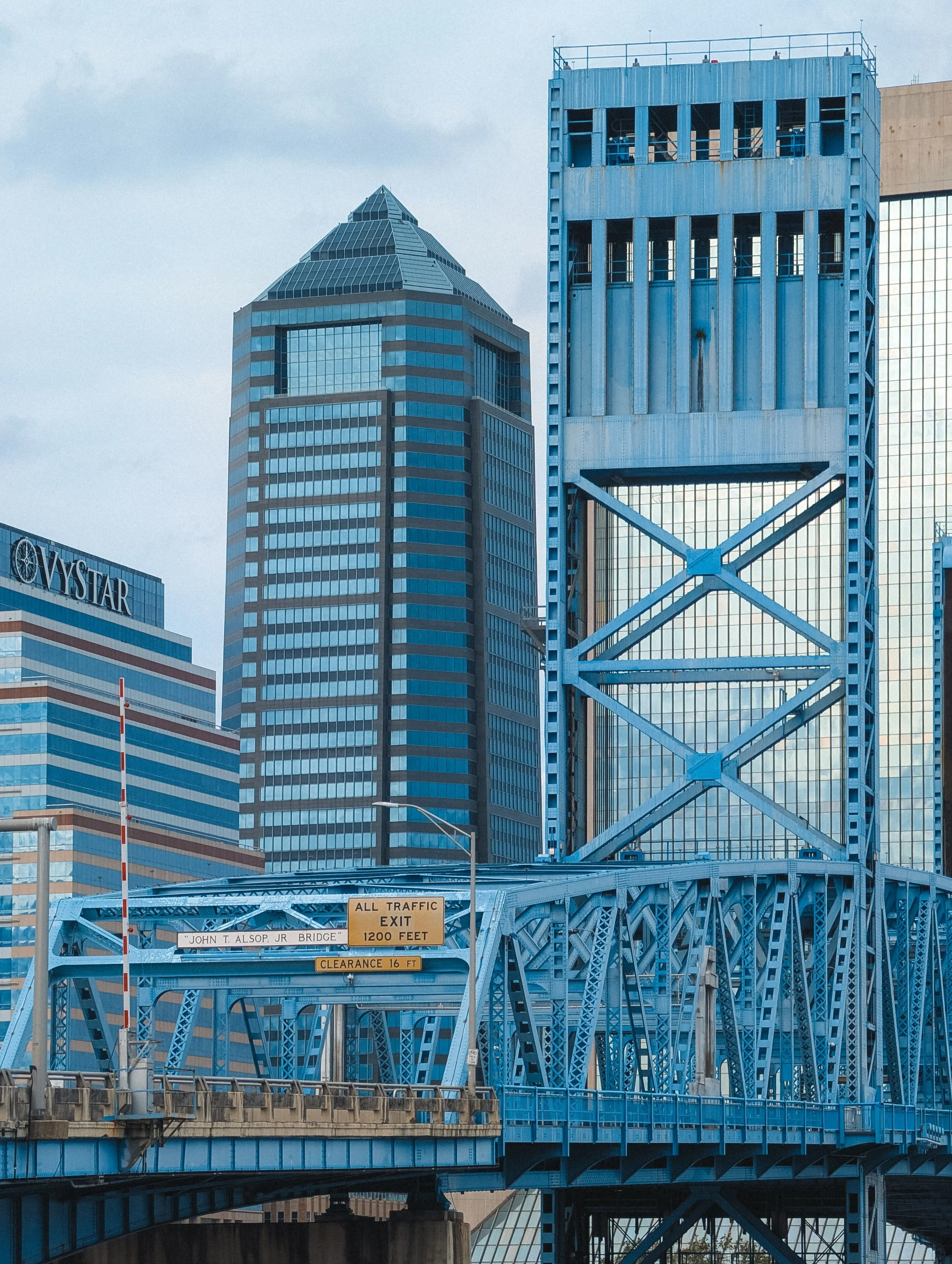 A blue steel bridge with signs indicating height restrictions and an exit, with tall modern buildings in the background.
