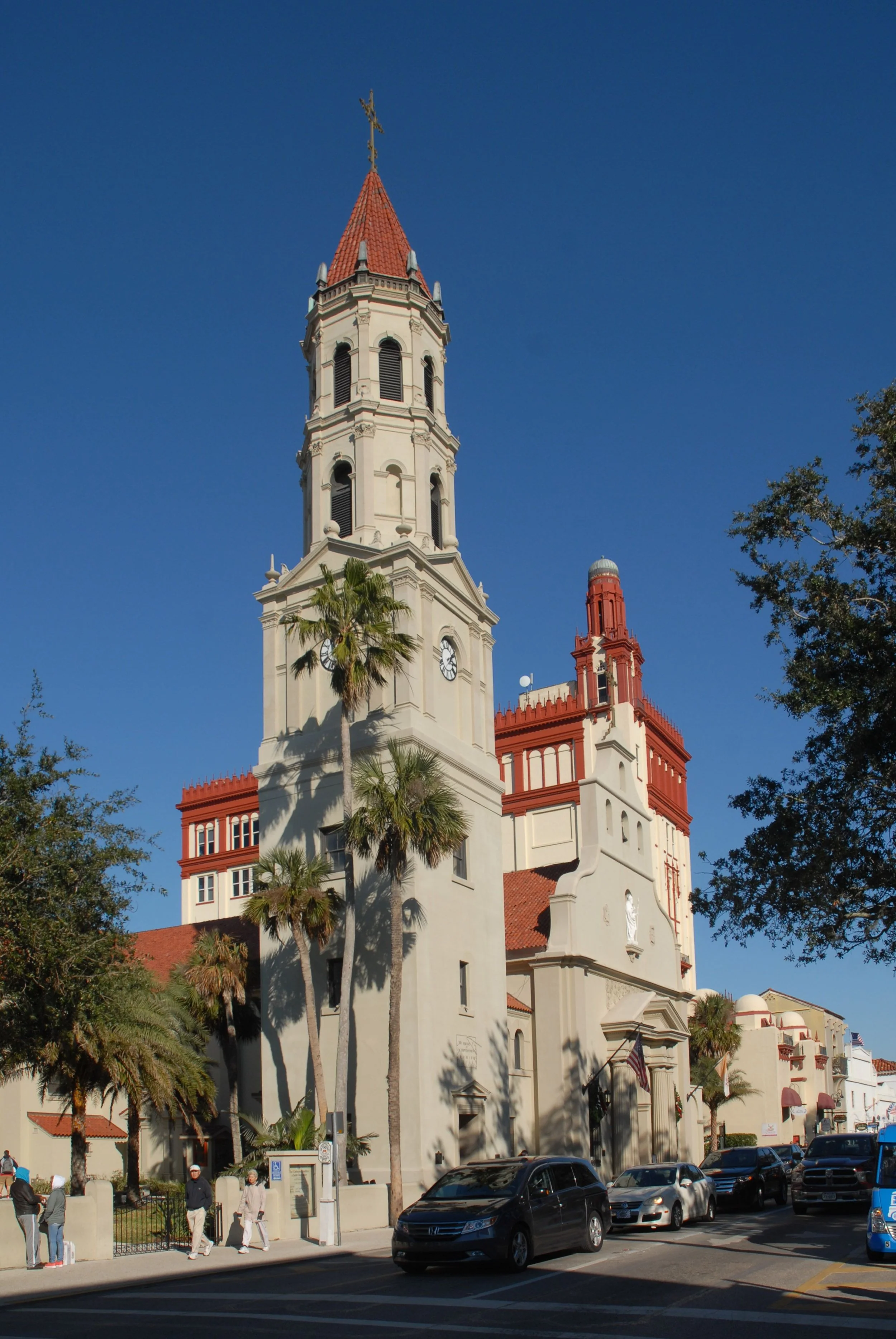 Historical church with a tall clock tower, red-tiled roof, and white walls, surrounded by palm trees, parked cars, and pedestrians on the street.