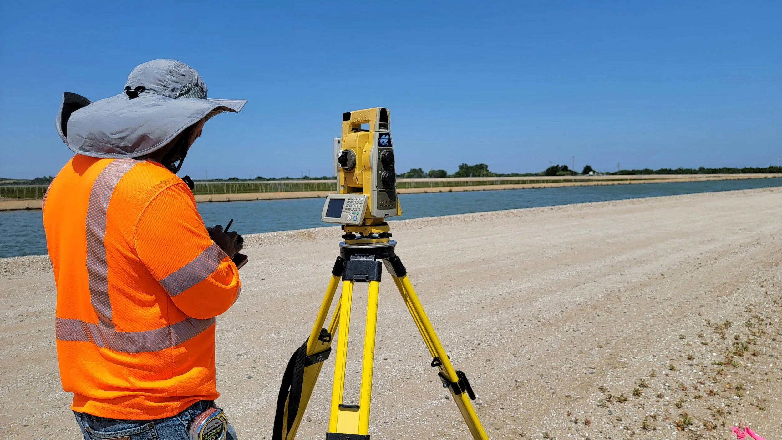 A surveyor wearing an orange safety vest and a wide-brimmed hat operating a theodolite on a tripod near a body of water under a clear blue sky.