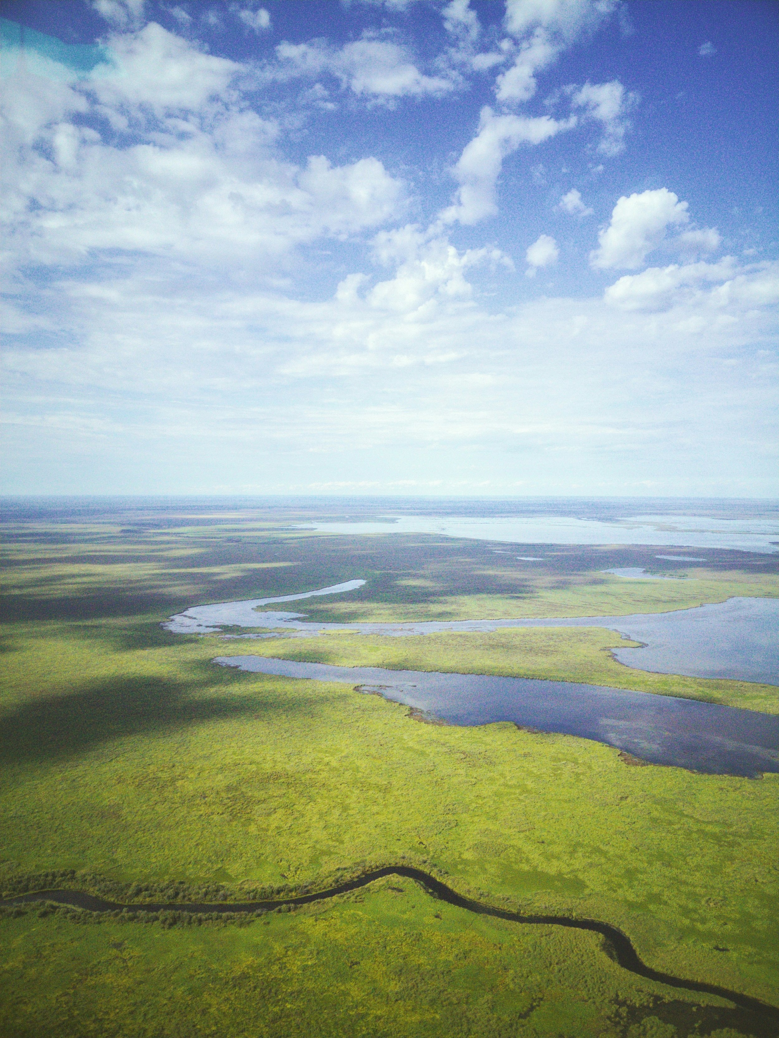 Aerial view of wetlands with patches of green vegetation, winding water channels, and a large body of water under a partly cloudy sky.