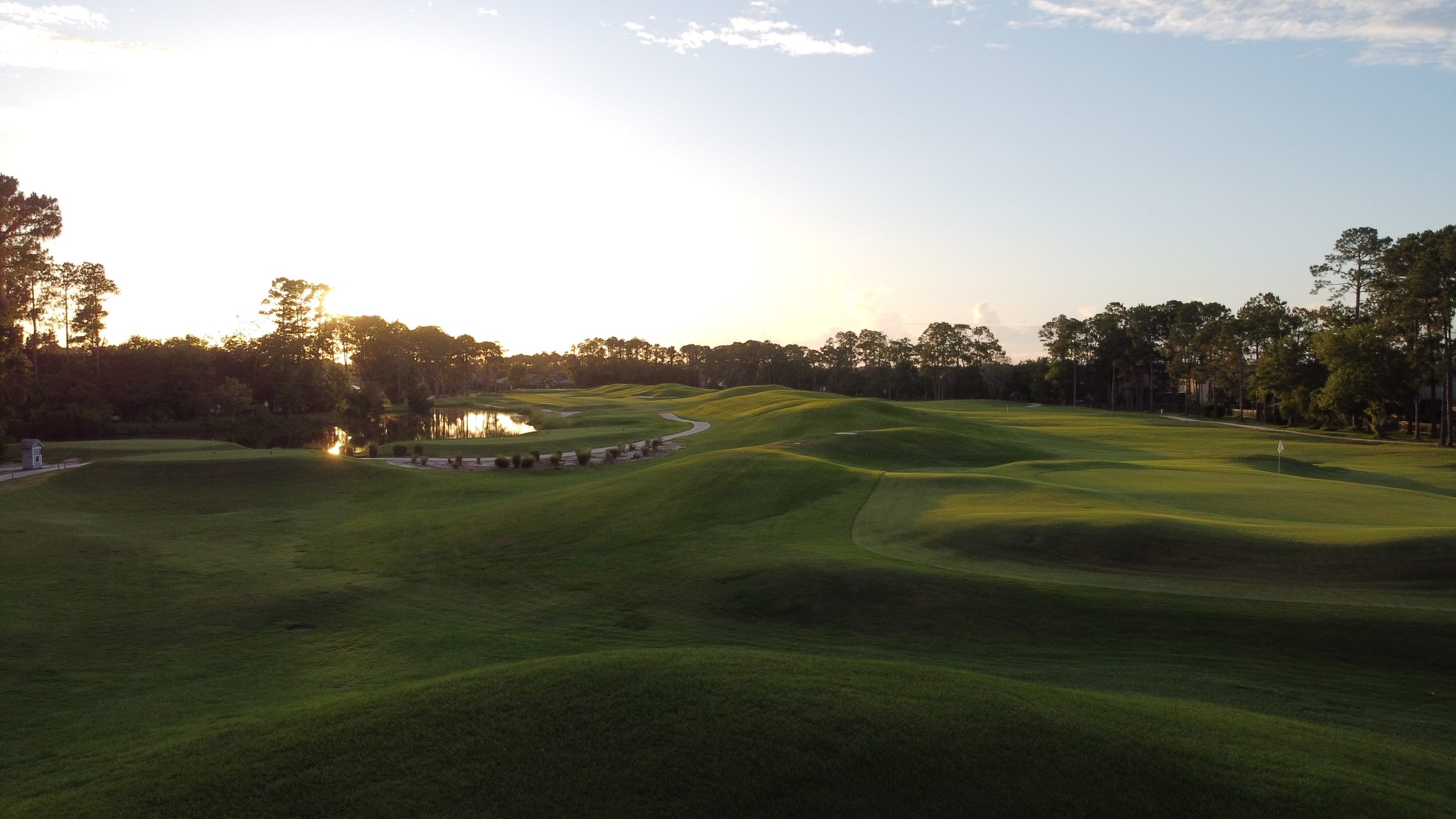 A golf course with lush green fairways, small water hazards, tall pine trees, and a flag on the putting green, at sunset.